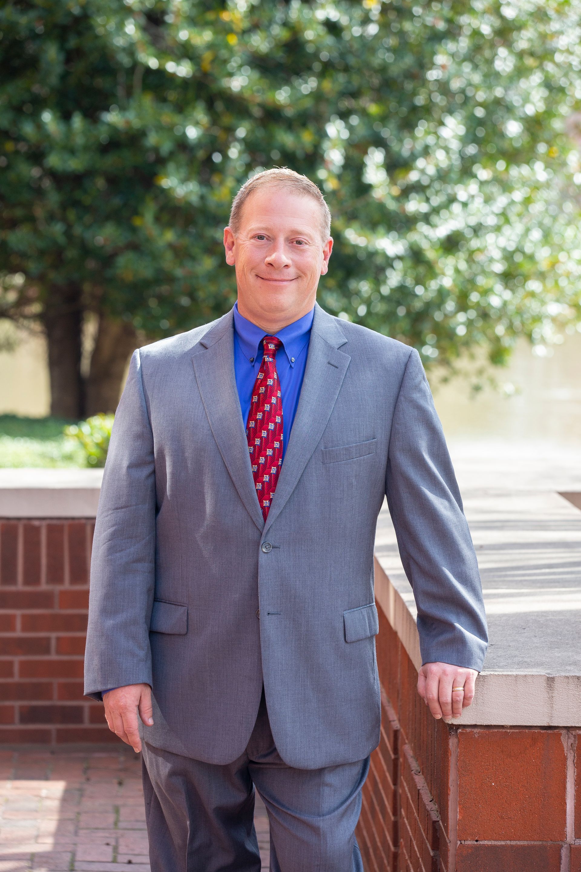 Man in a suit smiles outdoors; red tie, blue shirt, gray jacket, brick wall and greenery in background.