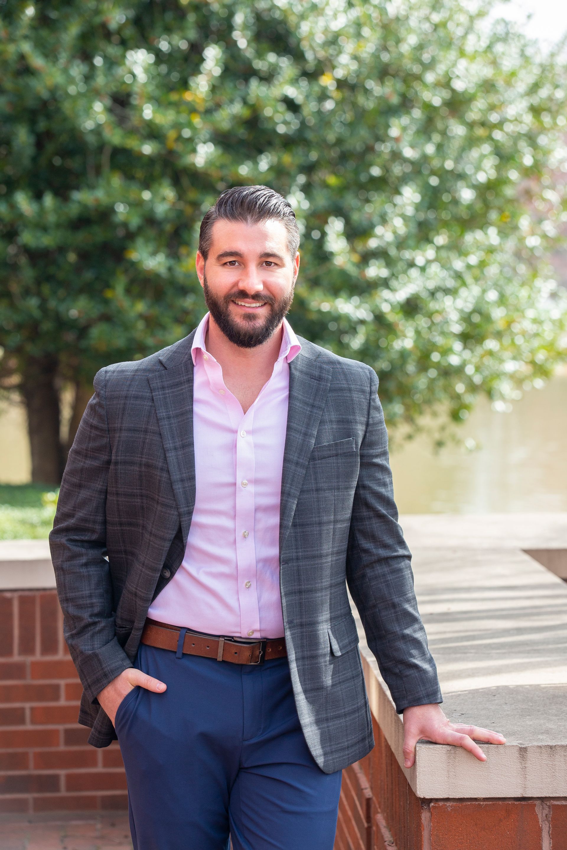 Man in blazer, pink shirt, and blue pants smiles outdoors with a hand in pocket.