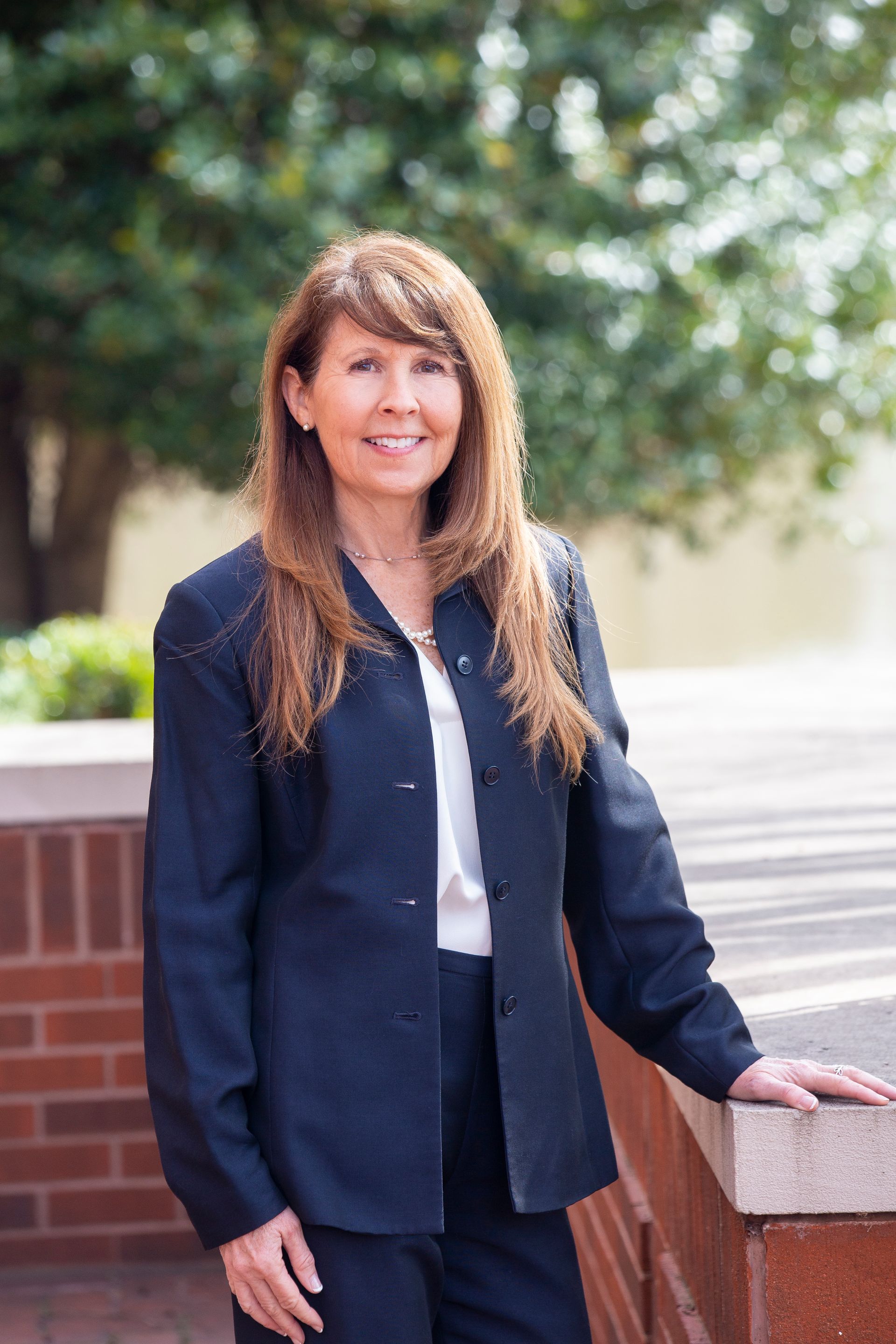Woman with long brown hair, wearing a navy blazer and white blouse, smiling outdoors.