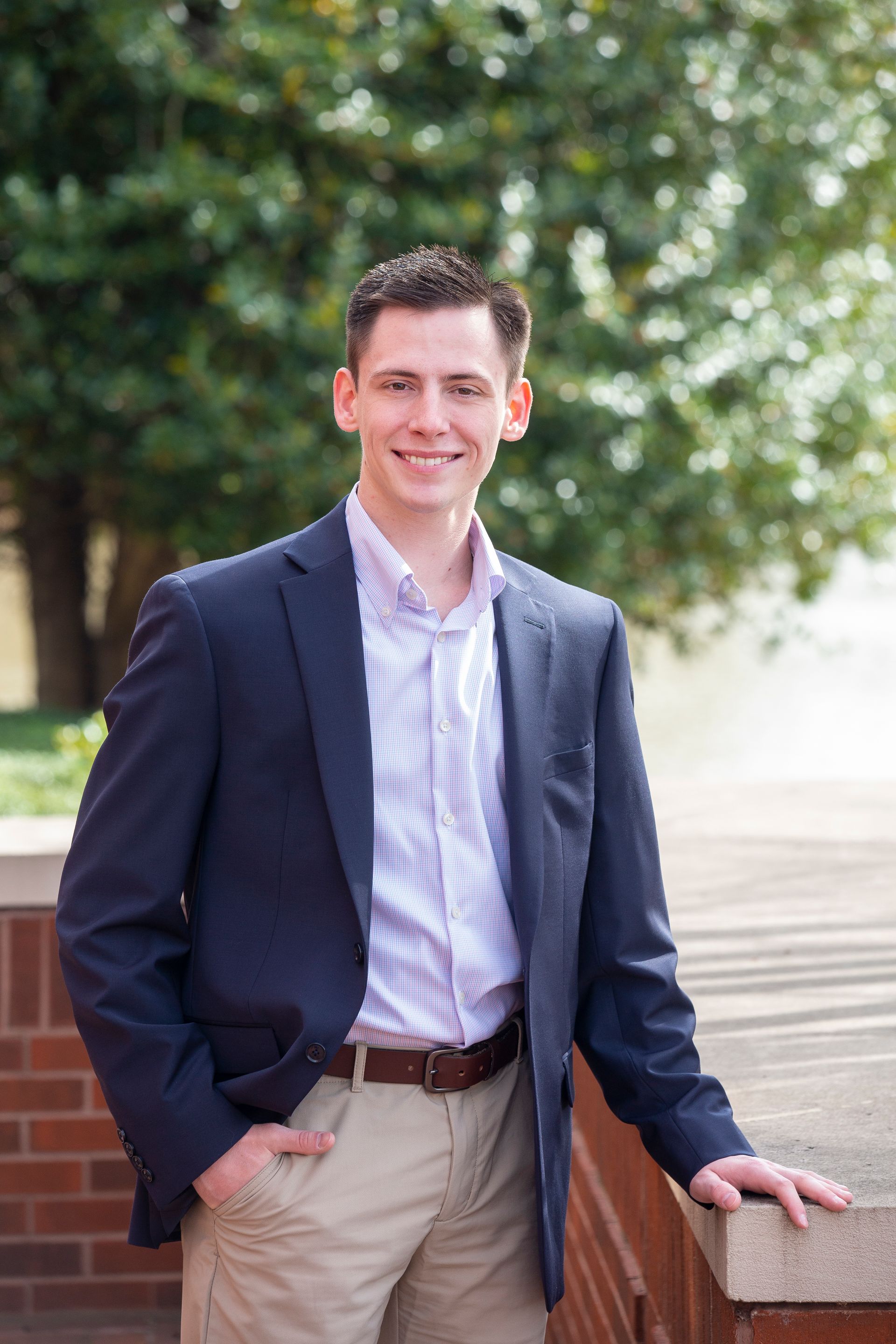Man in navy blazer and open-collar shirt smiles outside, trees in the background.
