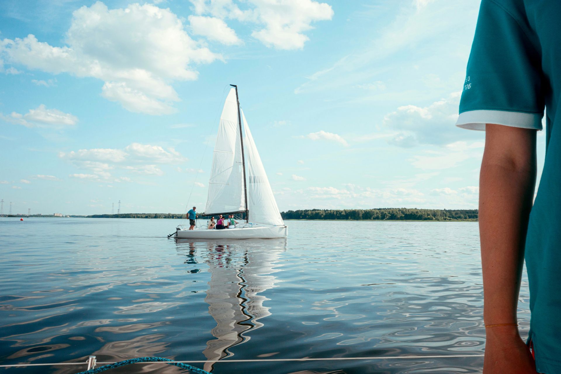 Sailboat on calm water, reflecting blue sky and clouds. Person in teal shirt in the foreground.