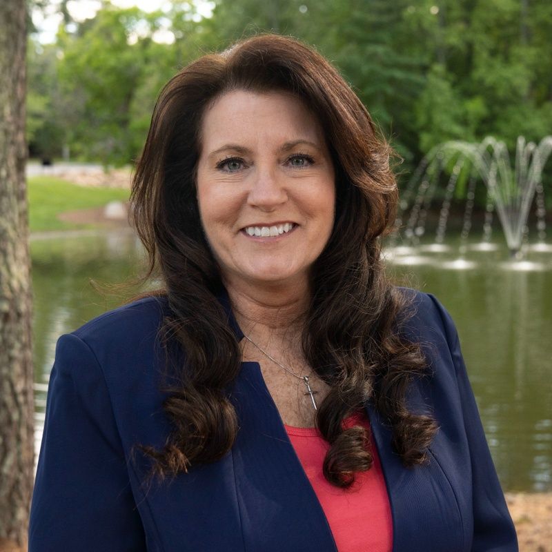 Woman with long brown hair smiling in a blue blazer, standing in front of a pond with a fountain.