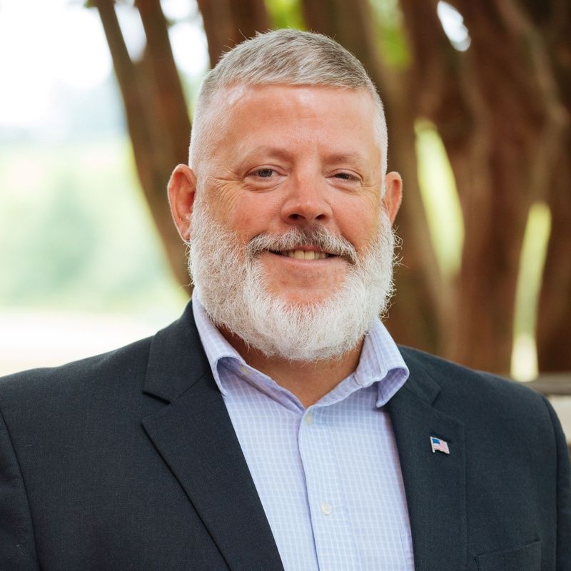 Man with graying beard, smiling in a suit, light blue shirt. Outdoor setting.