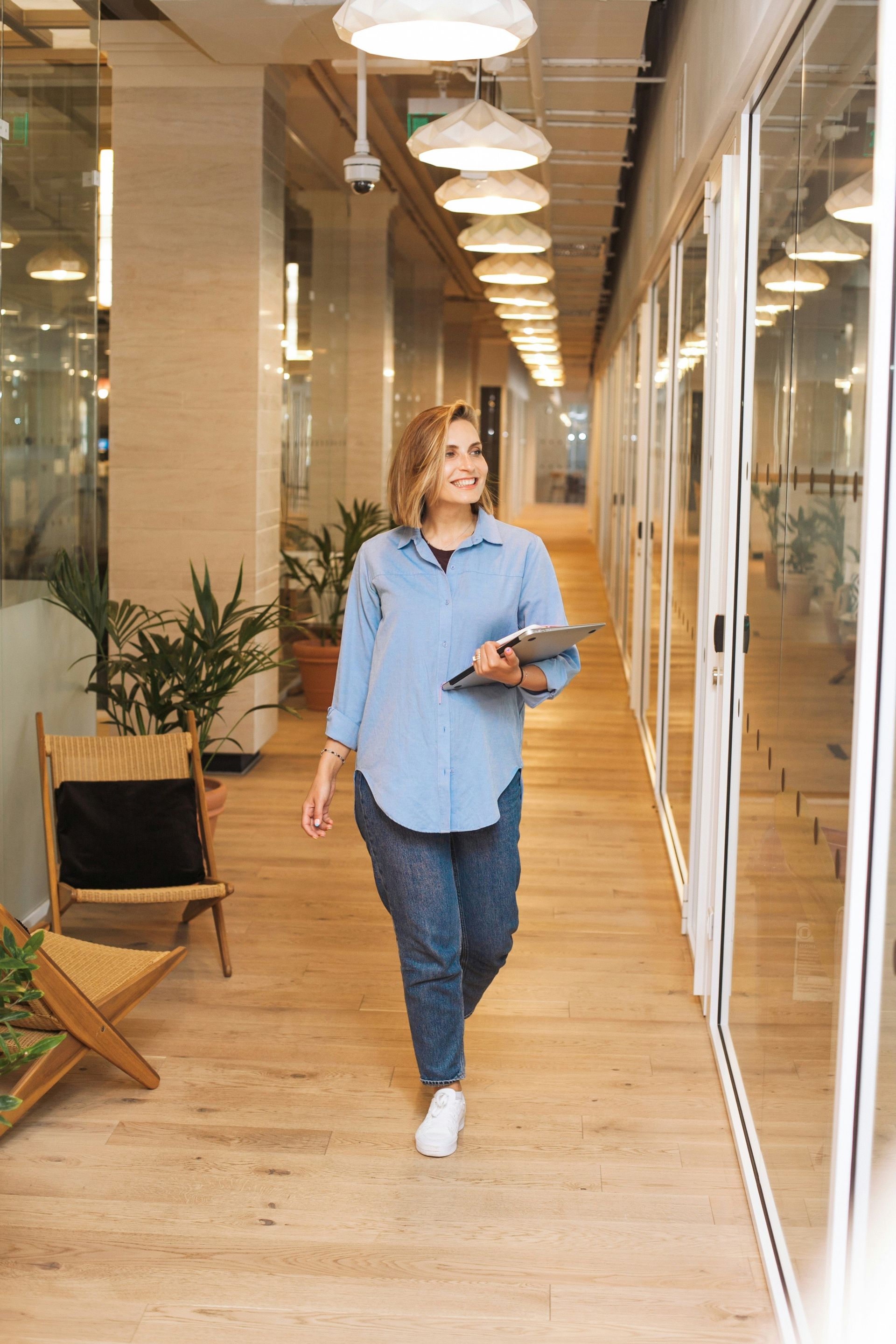 Woman smiling, walking down office hallway, holding tablet, wearing denim shirt and jeans, sunlight.