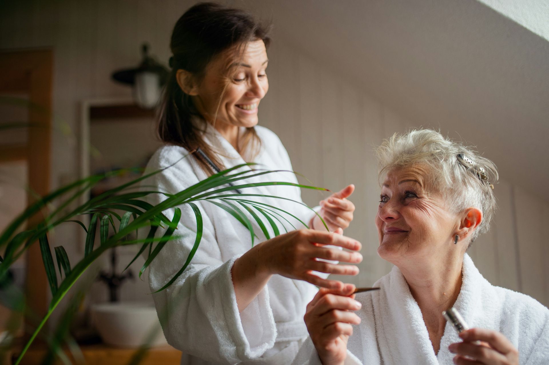 Woman applies makeup on older woman's face, both in white robes, smiling in a bright room.