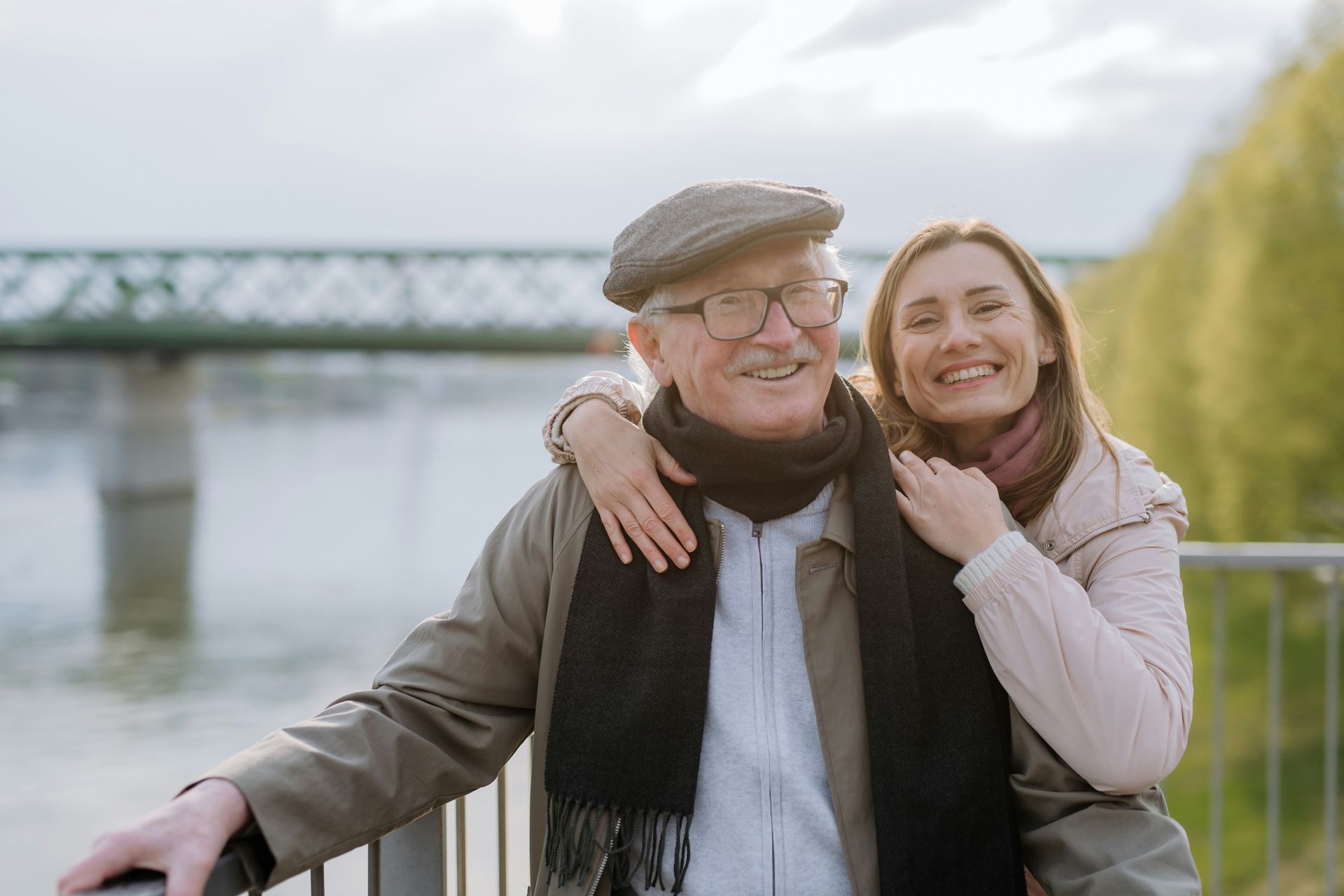 Elderly man with glasses and cap, and younger woman smiling, hugging near a bridge.