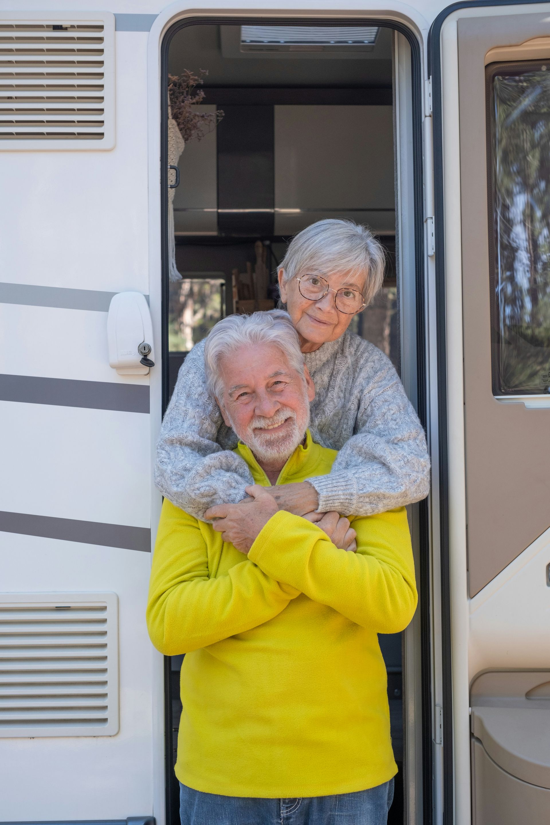 Elderly couple hugging in a RV doorway; man in yellow, woman in gray, smiling.