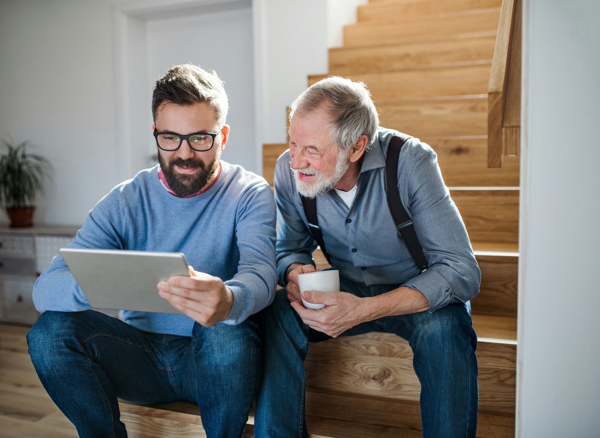 Man and older man, sitting on stairs, looking at a tablet, smiling. Bright, indoor setting.