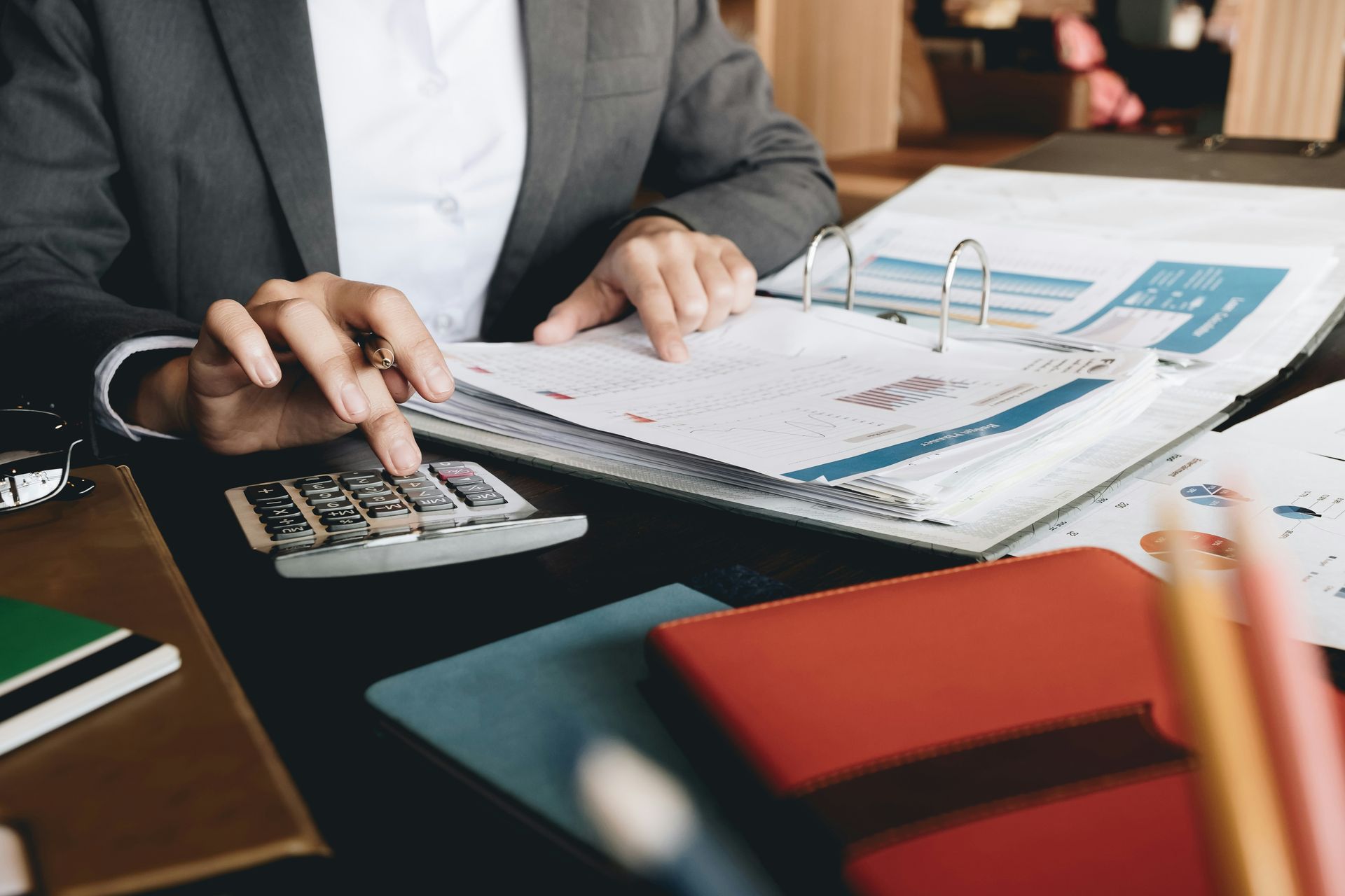 Person in a suit calculating finances with a calculator and papers in a binder on a desk.