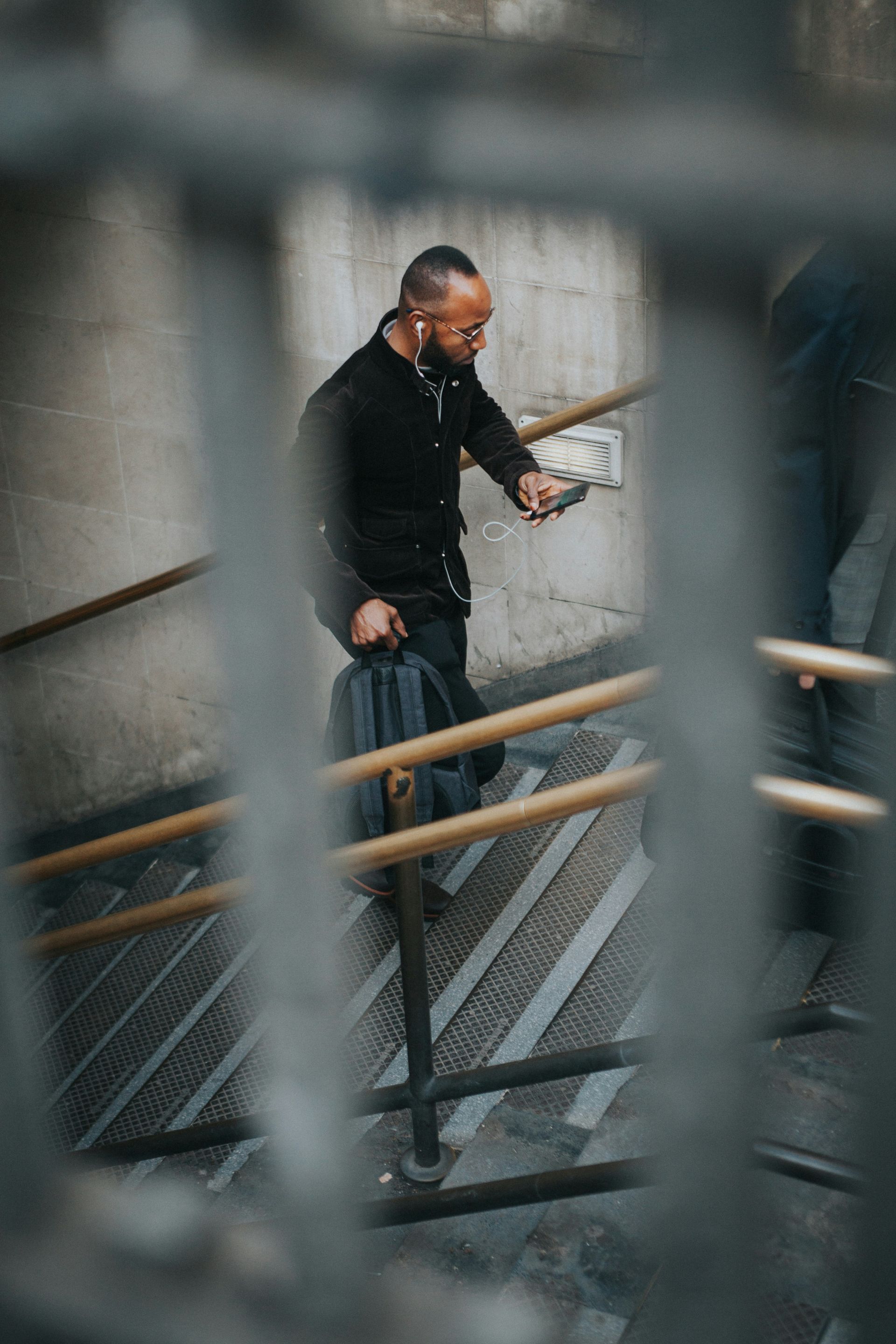 Man with backpack walking up stairs, looking at his phone through a metal gate.