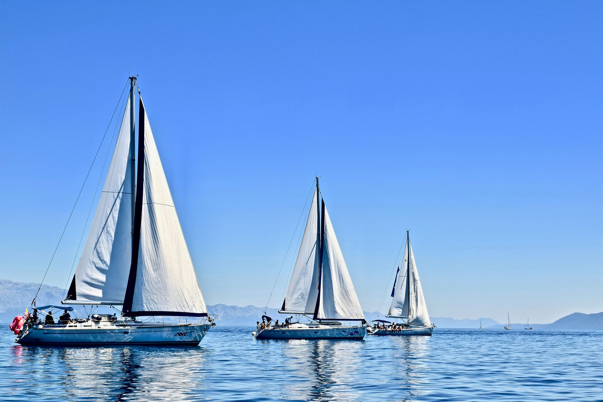 Three sailboats with white sails on a blue ocean under a clear blue sky.