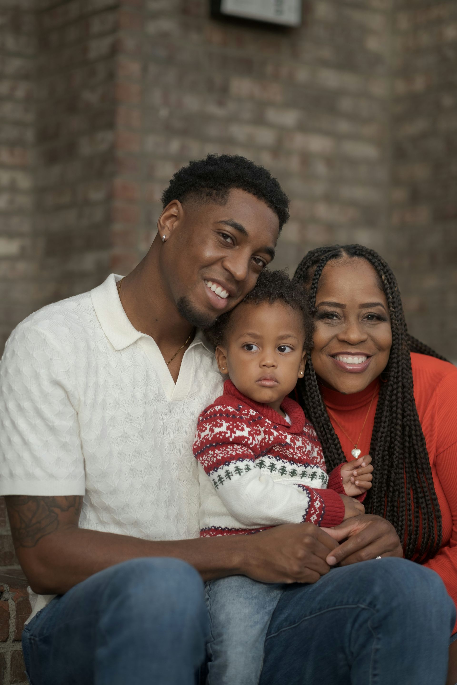 Family of three smiling for the camera. Man in sweater, woman in red, holding child in sweater. Against brick.