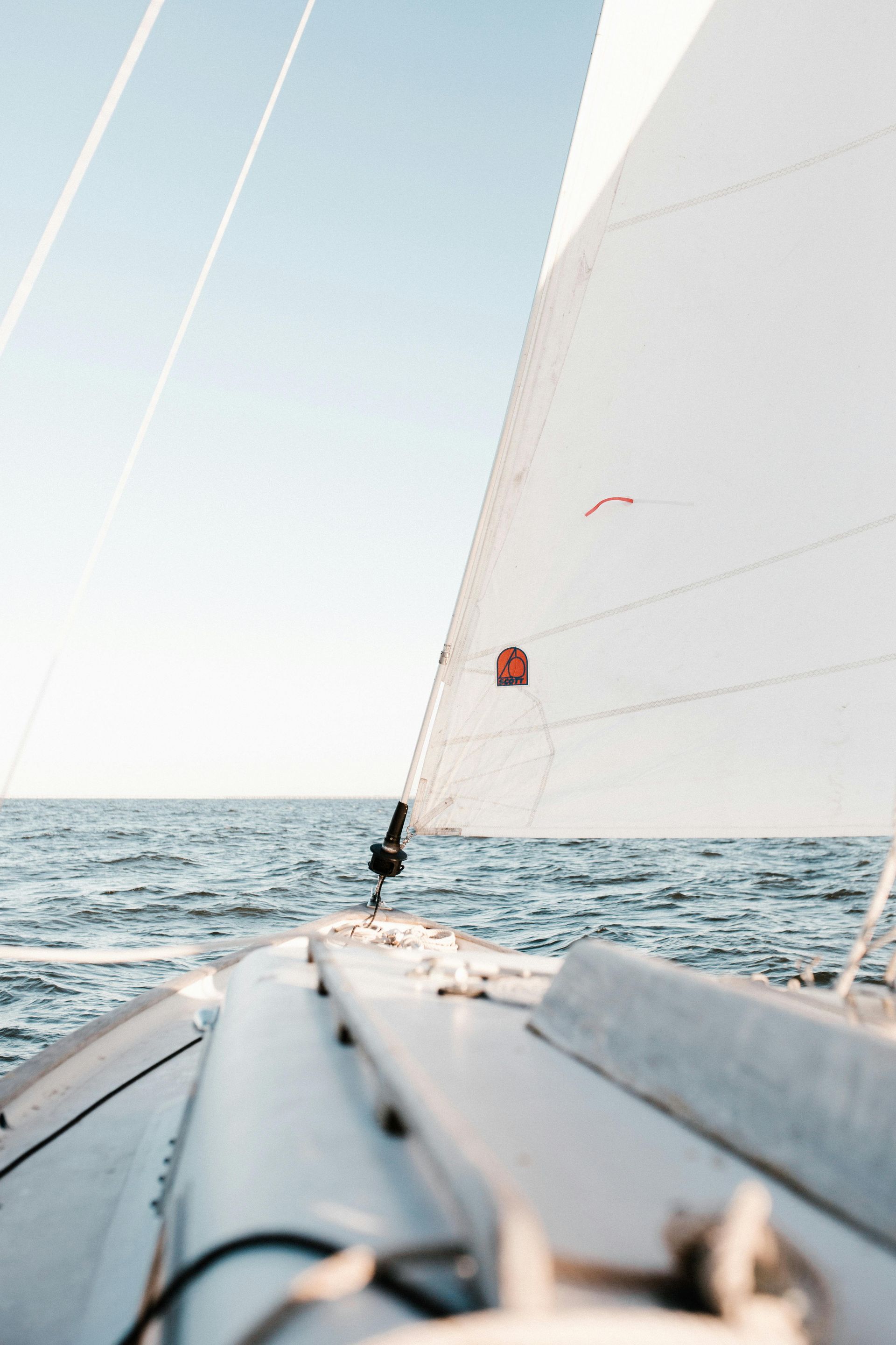 Sailboat sailing on a calm blue sea, with a white sail against a light blue sky.