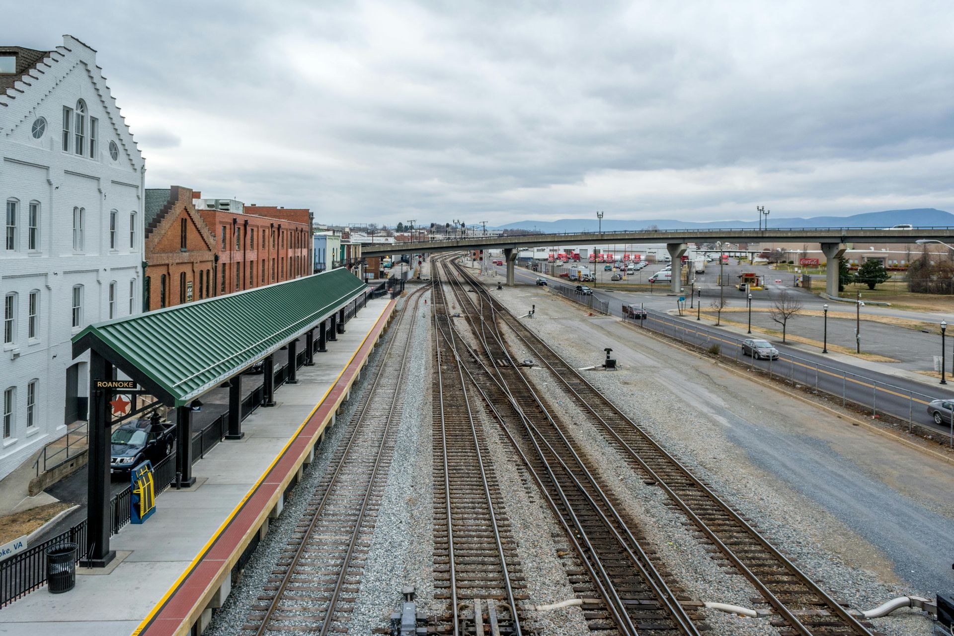 Train tracks with a covered platform, buildings, and overpass under cloudy skies.