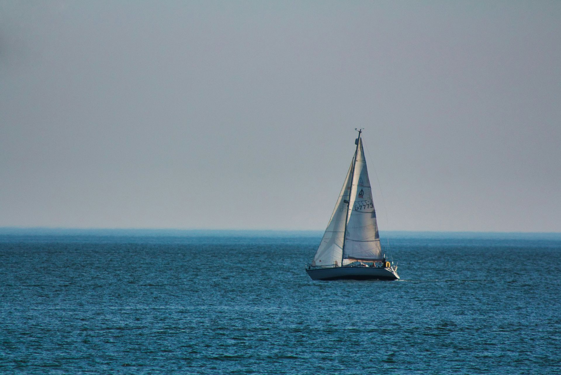 Sailboat sailing on a blue sea under a light gray sky.