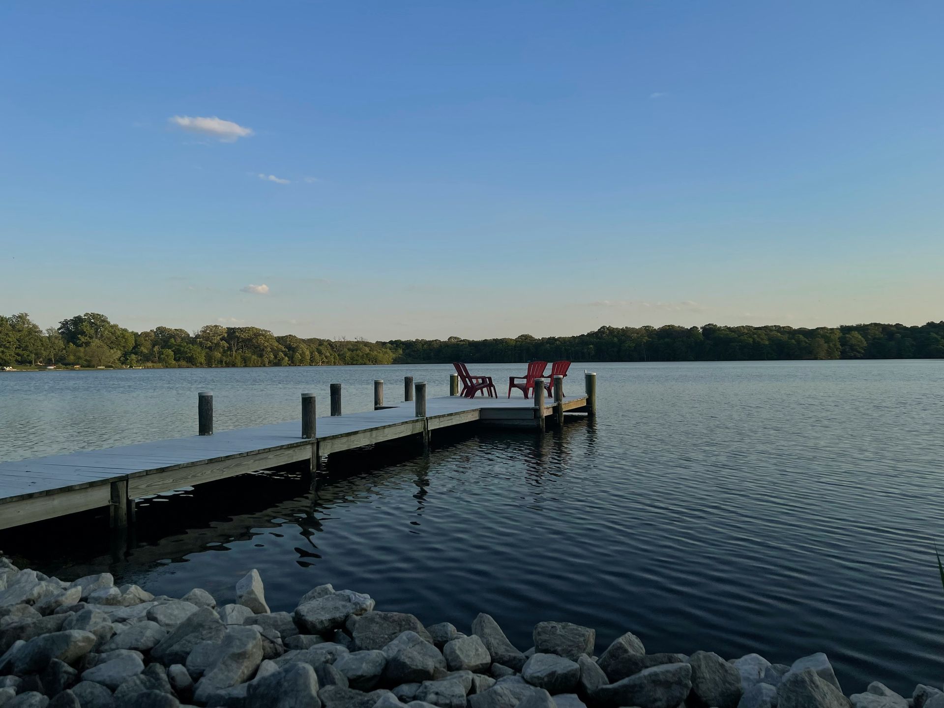 Dock with red chairs on a calm lake under a blue sky; trees line the opposite shore.