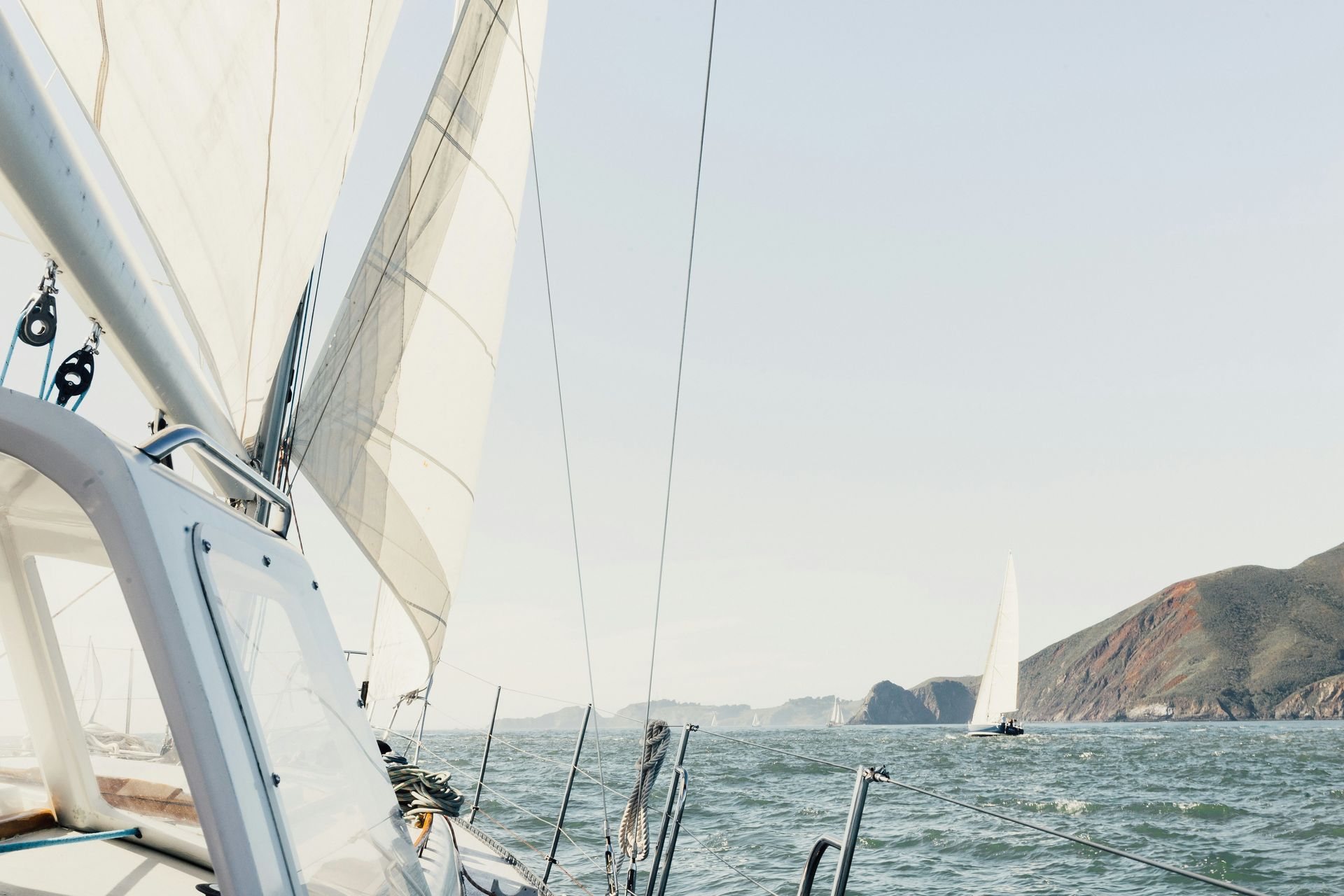 Sailboat on water, sails up. Another boat and land in the distance on a sunny day.