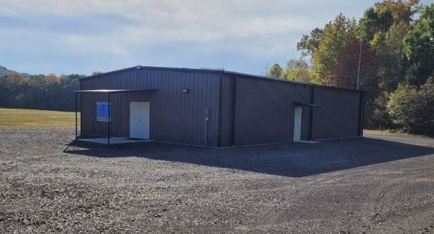 Brown metal building with white doors and a small porch on a gravel lot; trees in background.