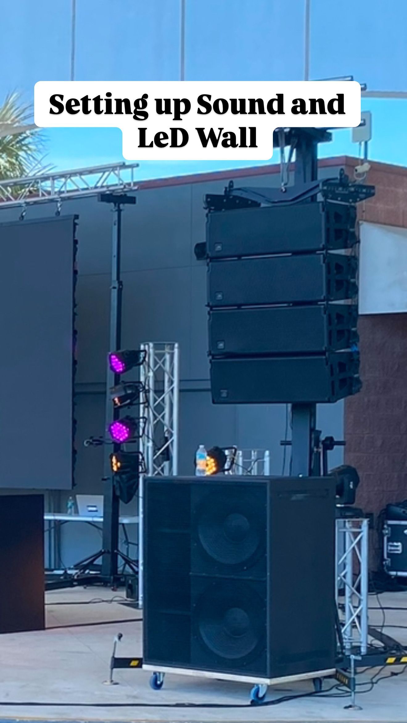 Sound equipment setup with a line array speaker stack, a large subwoofer on wheels, and an LED screen under a clear sky.
