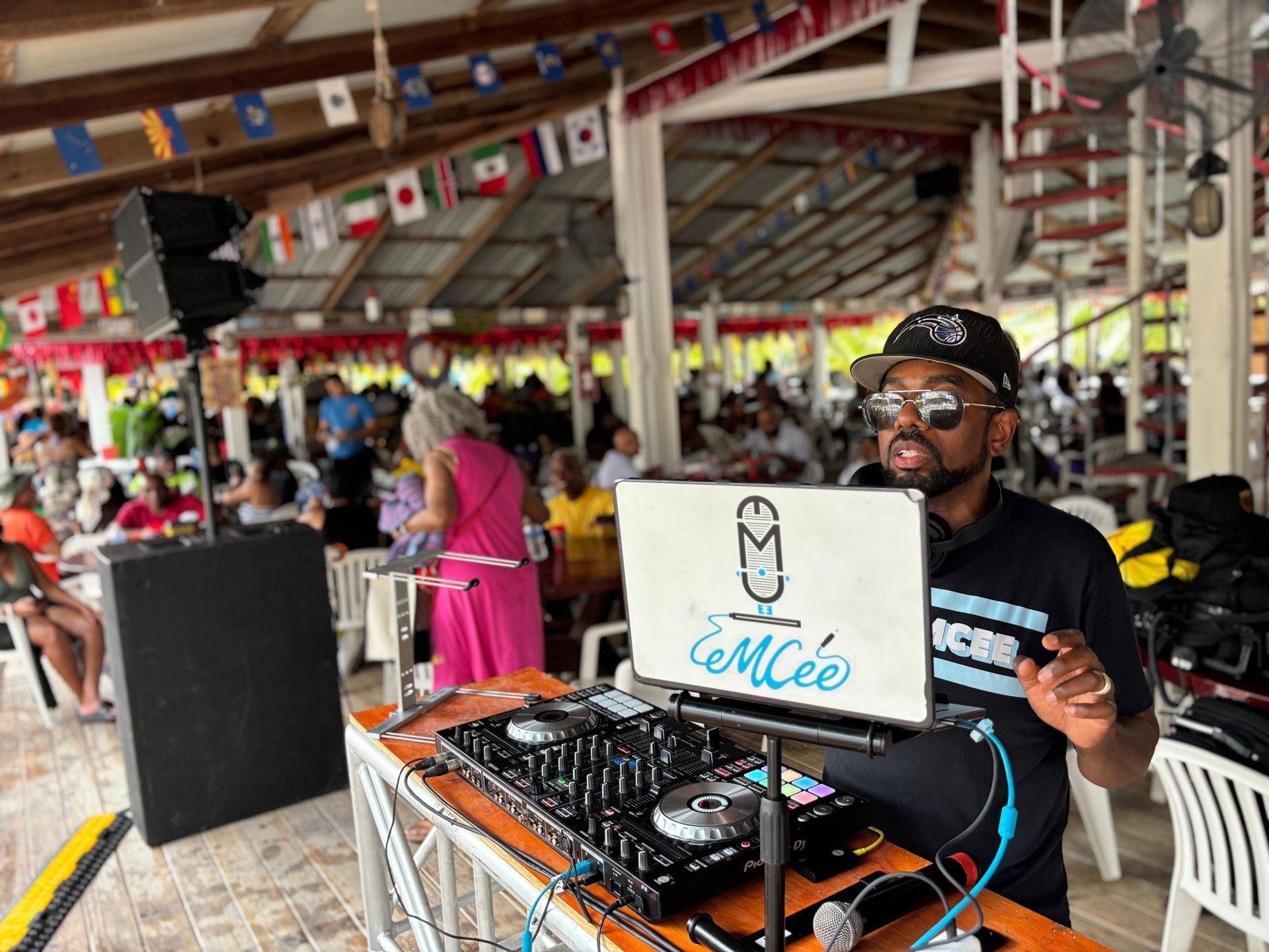 A DJ in a black cap and shirt mixes music on a console in a bustling, open-air venue decorated with international flags.