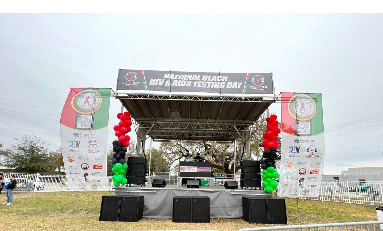 An outdoor stage for a National HIV & AIDS Testing Day event, decorated with red, black, and green balloon pillars.