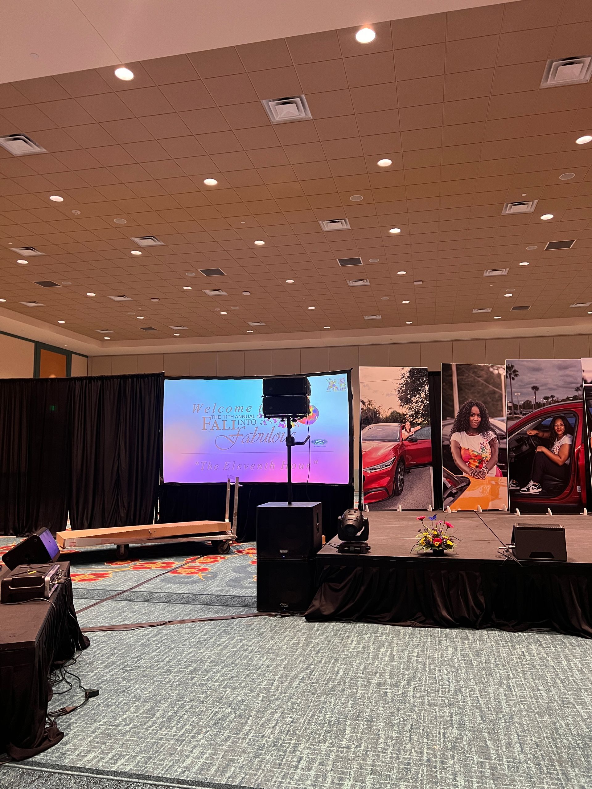 A stage setup in a hotel ballroom with a digital screen, black curtains, speakers, and large printed banners.
