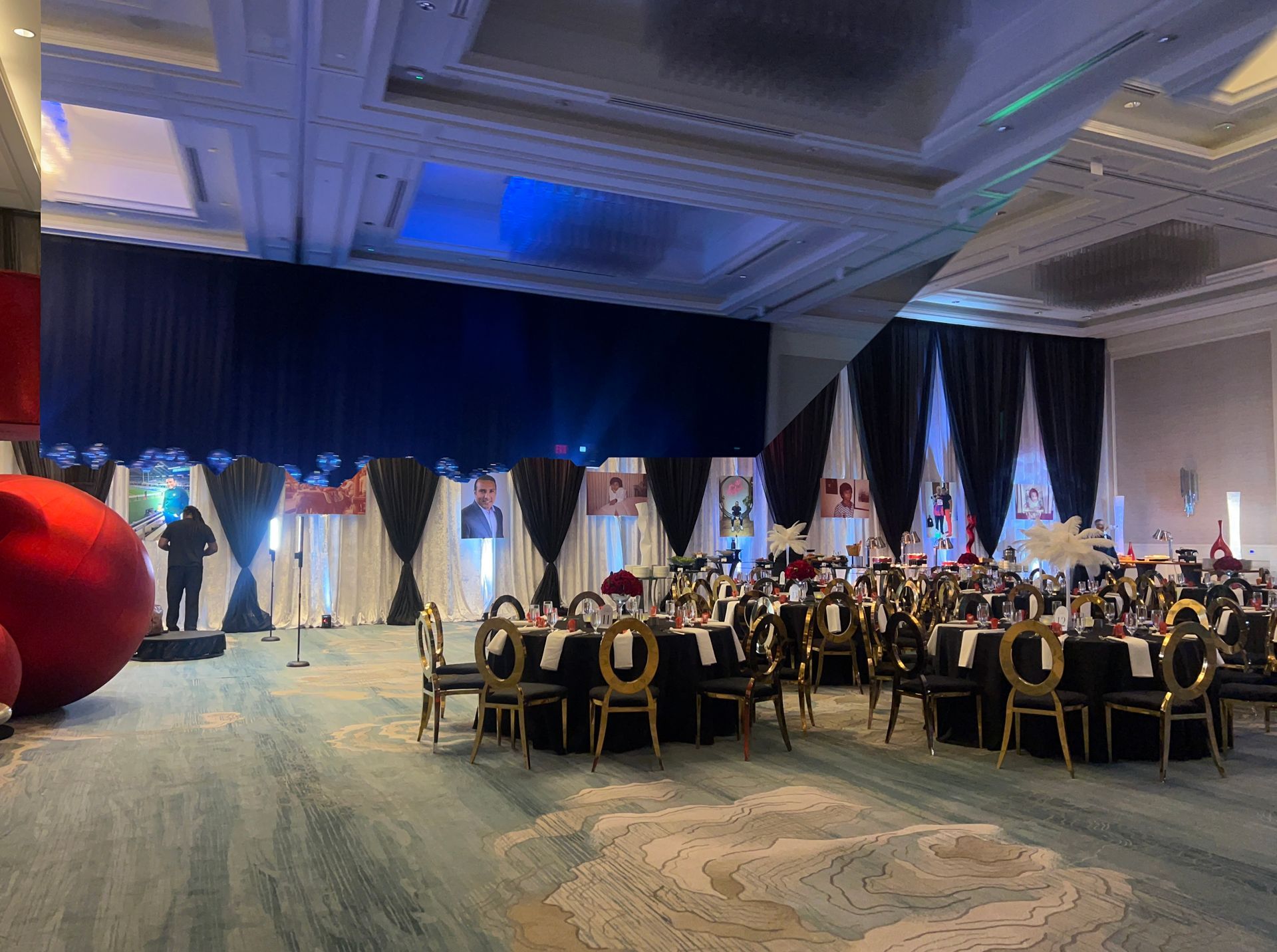 A banquet hall set for a formal event with round tables, gold chairs, and a stage decorated with white and black drapes.