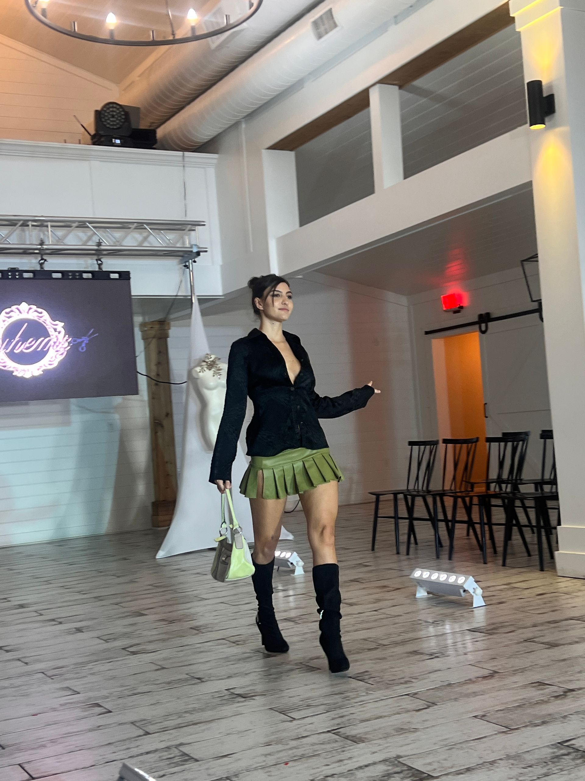 A model walks down a runway wearing a black blazer, a pleated olive green miniskirt, and black boots, holding a bag.