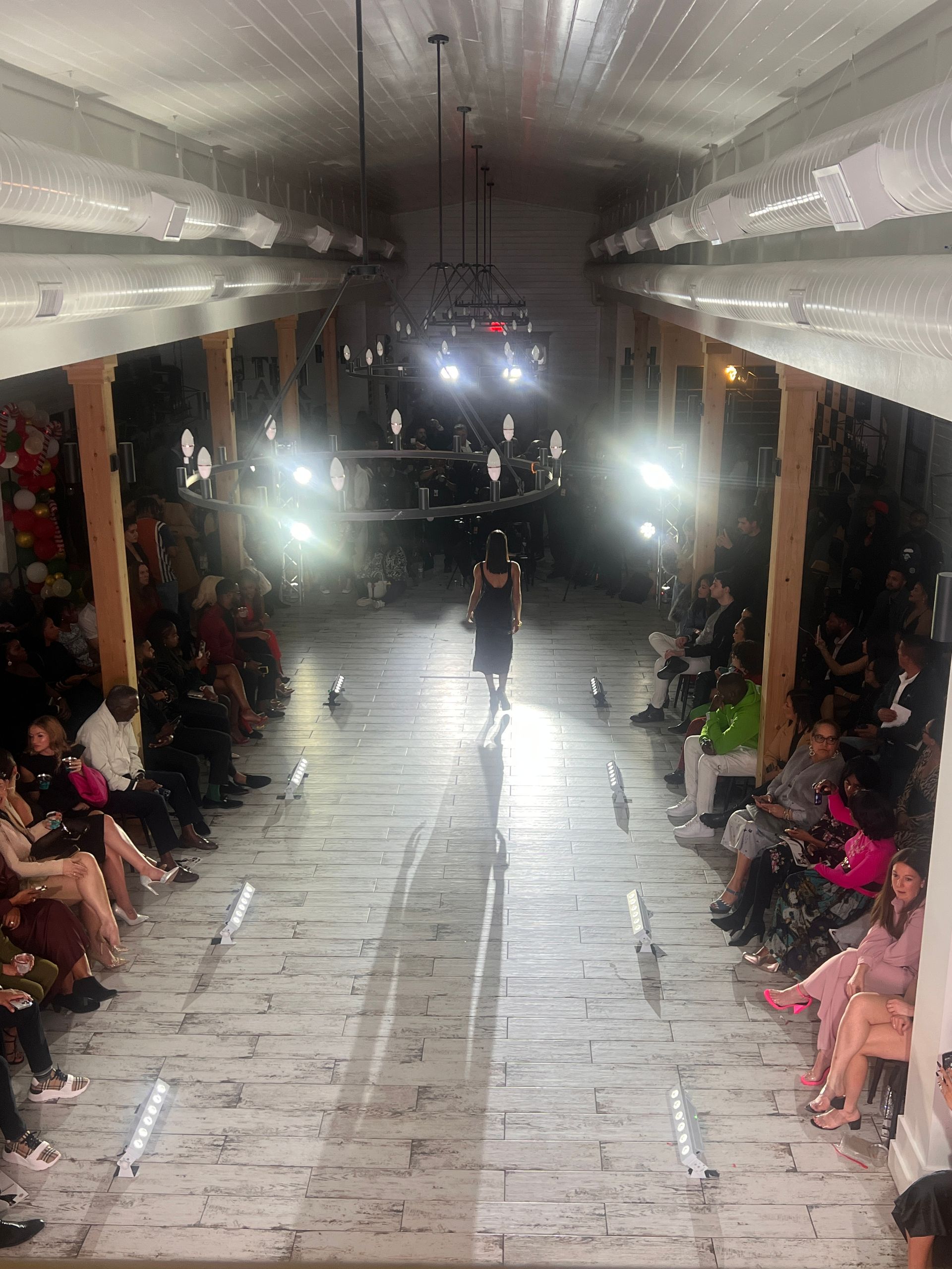 A fashion model walks down a rustic, dimly lit runway surrounded by seated audience members in a long, wooden venue.