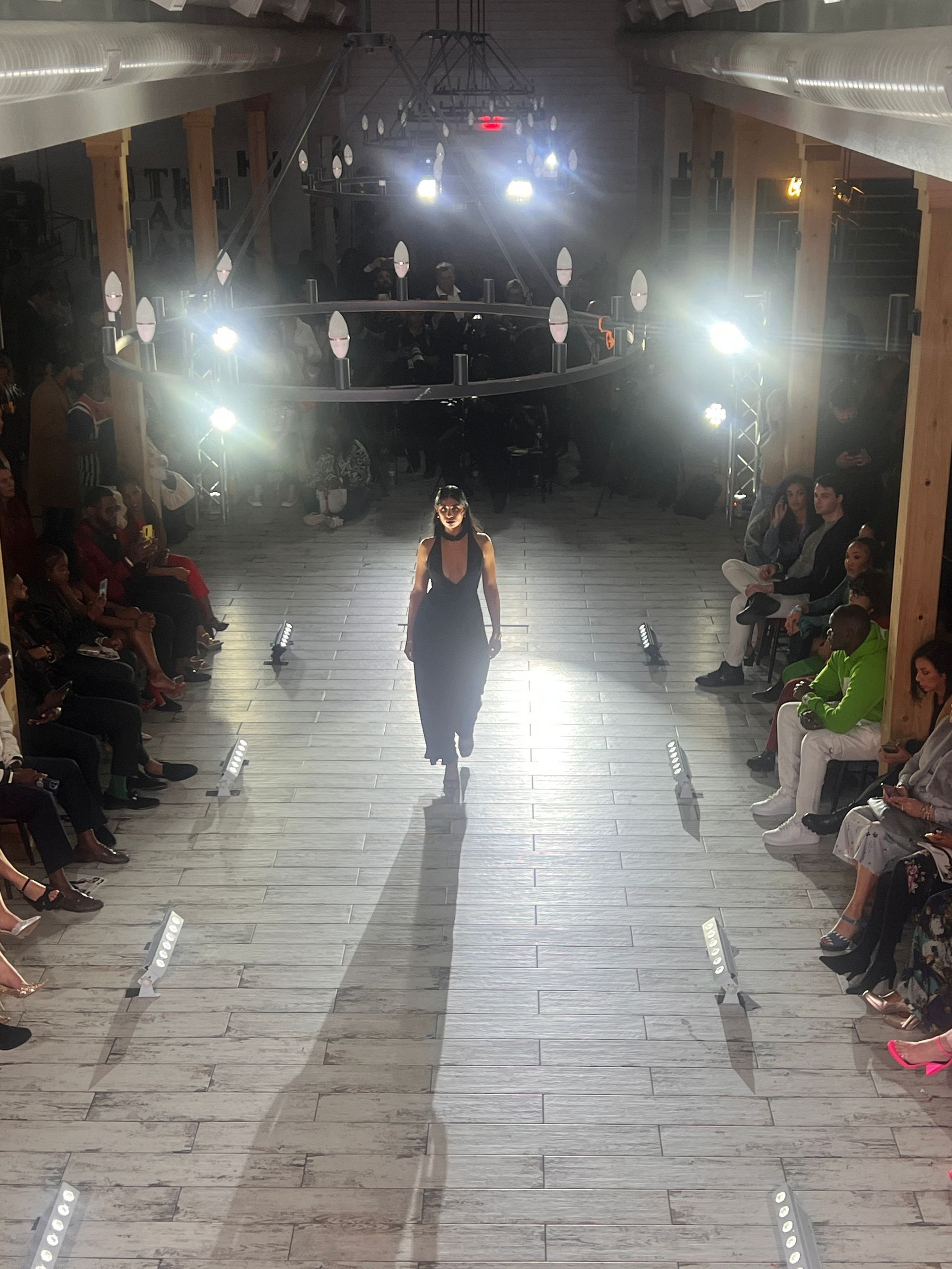 A model walks down a runway in a dimly lit, narrow venue, illuminated by bright overhead lights. Audience sits on sides.