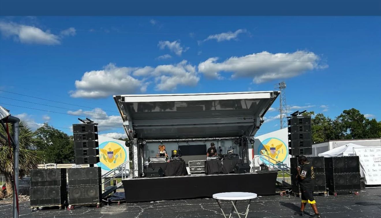 Outdoor stage setup with sound equipment and speakers under a blue sky with scattered clouds.