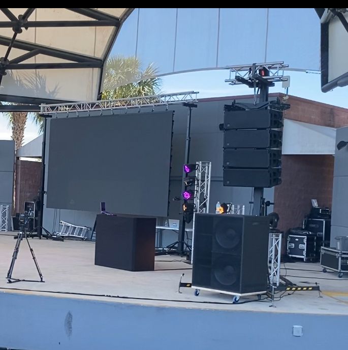 An outdoor concert stage with a large LED screen, a professional sound system, and lighting rigs on a paved surface.