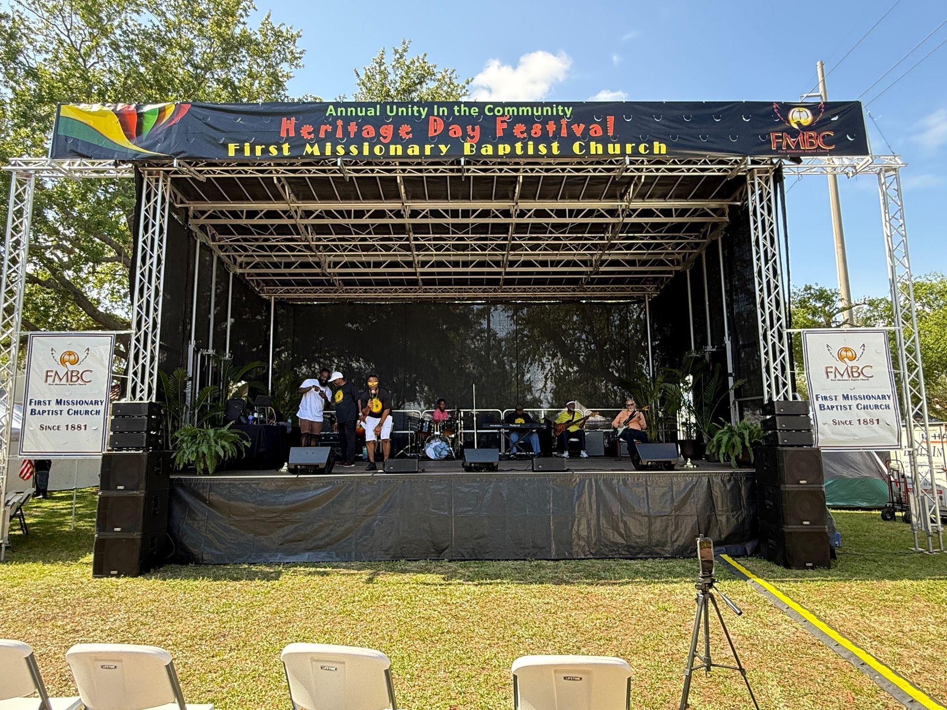A large stage at a Heritage Day Festival for First Missionary Baptist Church, with people sitting under a truss canopy.