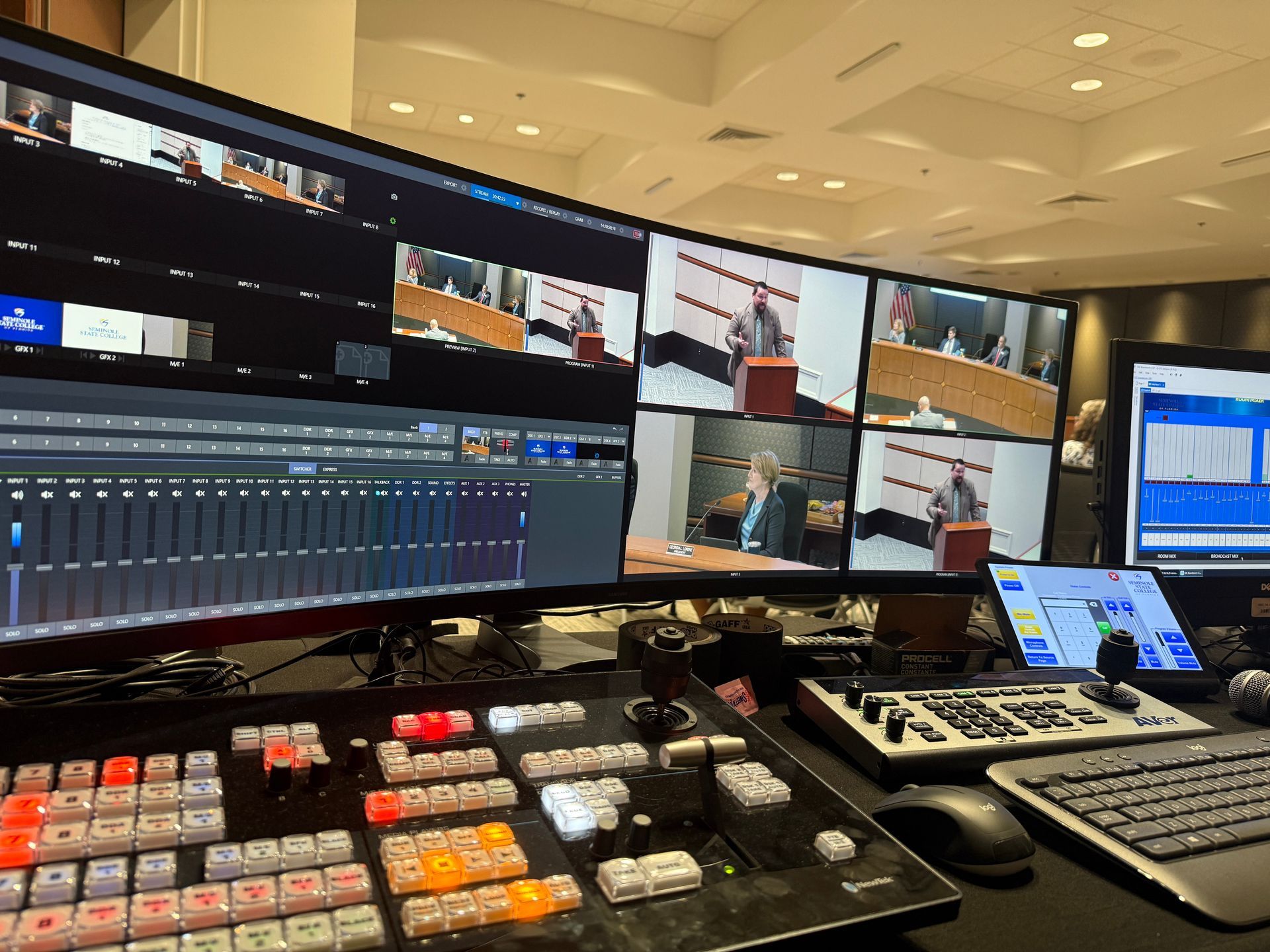 A broadcast control room showing multi-screen video feeds of a public meeting alongside a video switcher console.