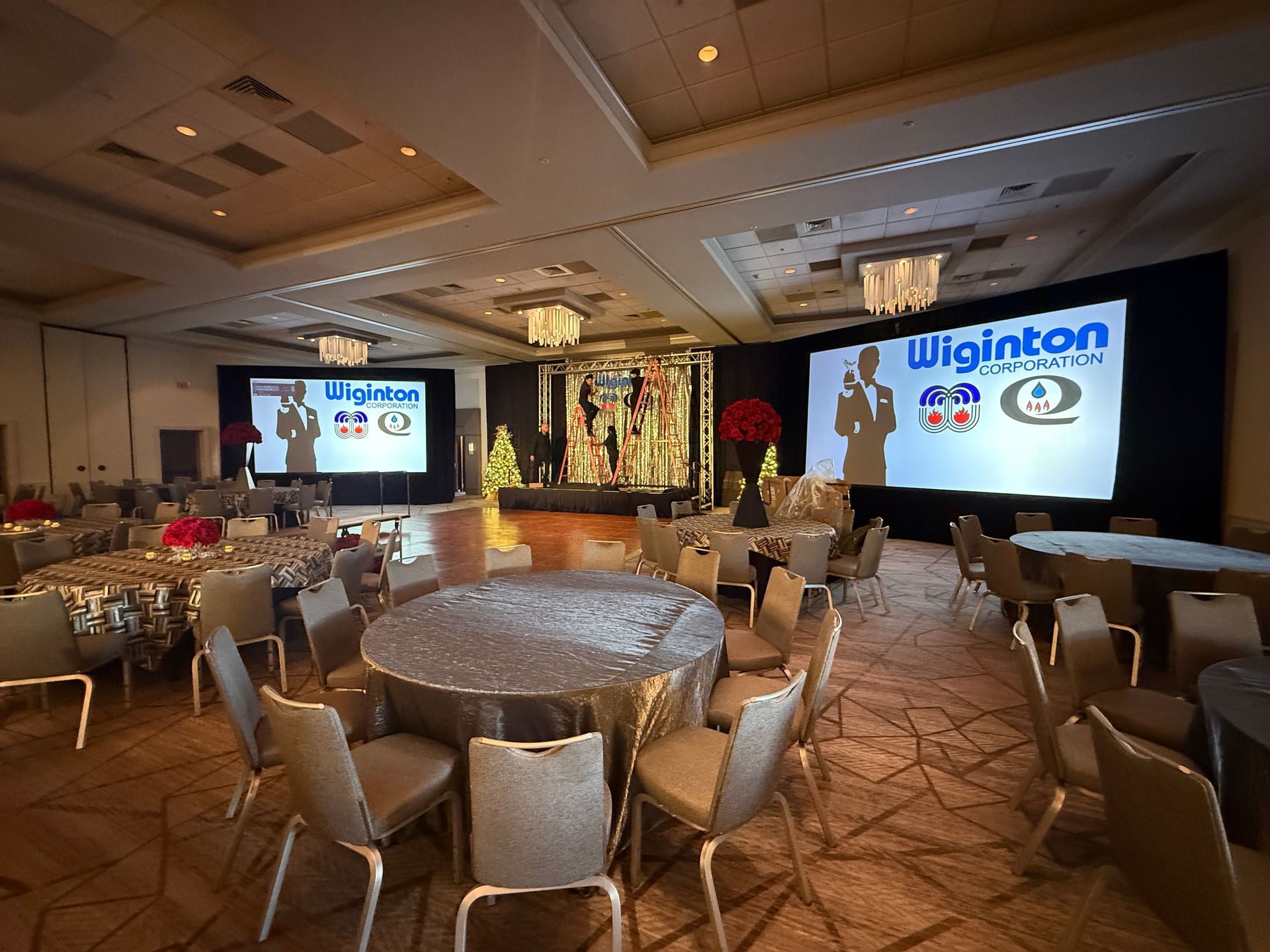 A banquet hall with round tables set for a Wiginton Fire corporate event, featuring large screens displaying branding.