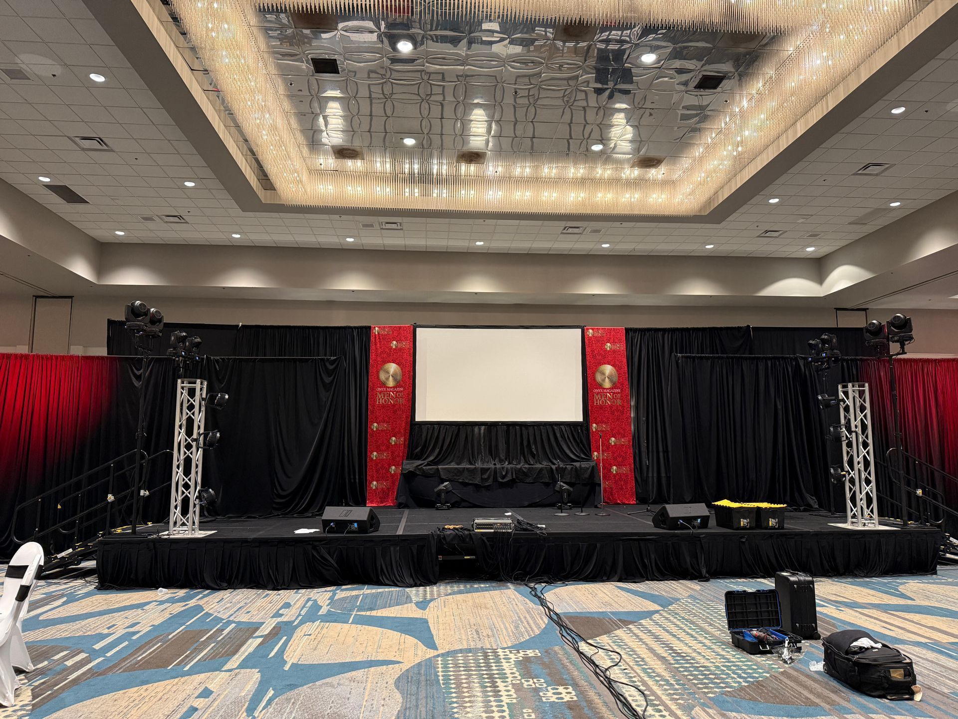 A stage setup in a ballroom featuring a center projection screen, black curtains, red branding panels, and metal trusses.