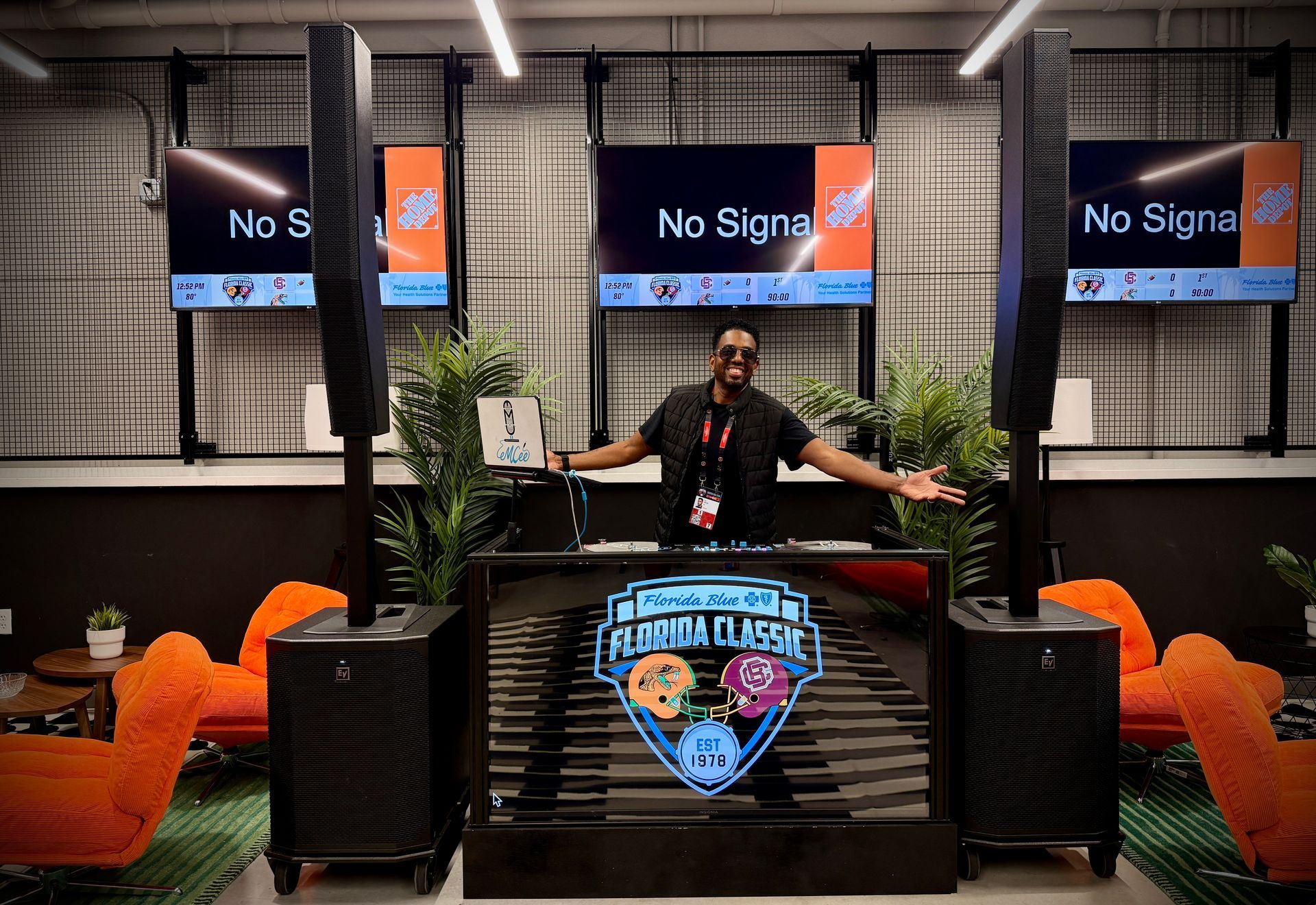 A person behind a DJ booth at the Florida Classic, with large speakers on both sides and orange chairs in a modern room.