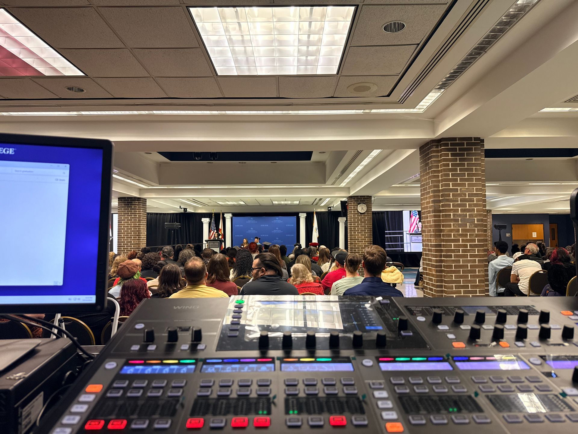 A view from a soundboard showing a crowd of people in a large hall with a stage, lighting, and decorative brick columns.