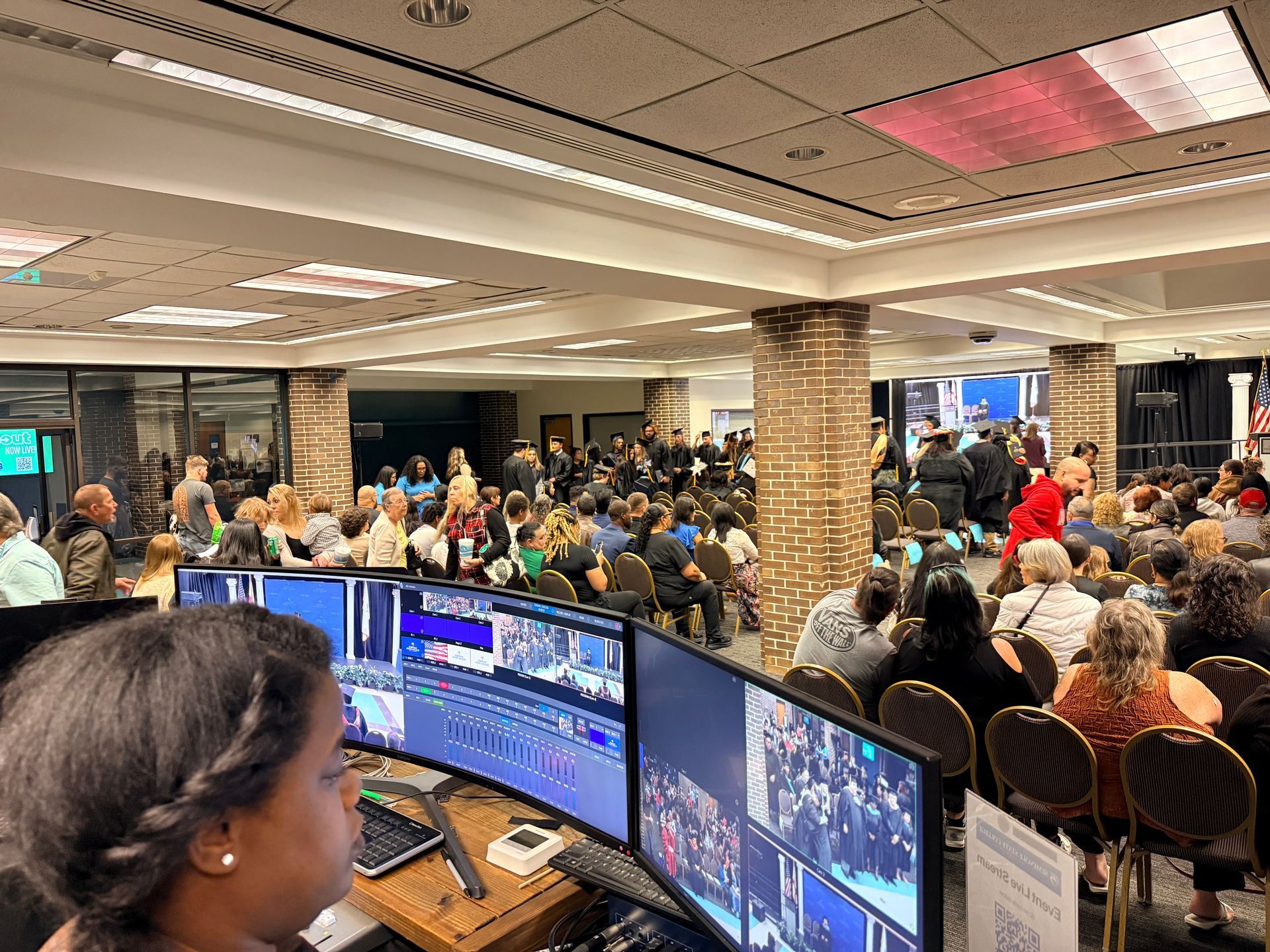 A person at a computer workstation monitors a video feed of a large group gathering in a brightly lit indoor venue.