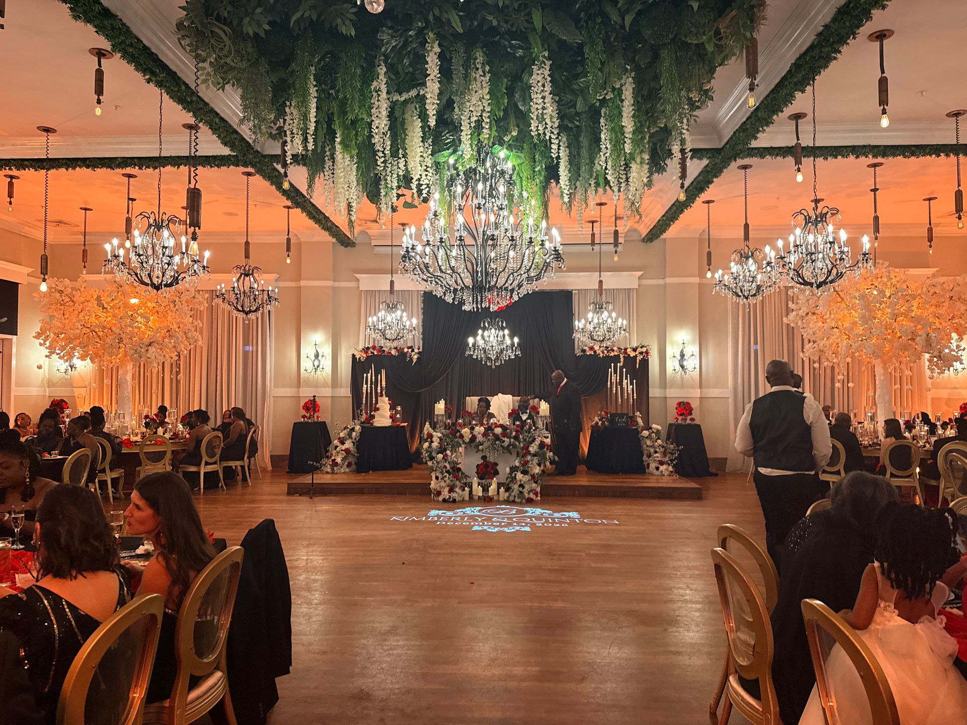 A banquet hall decorated with lush greenery, crystal chandeliers, and warm lighting, set for a formal event.