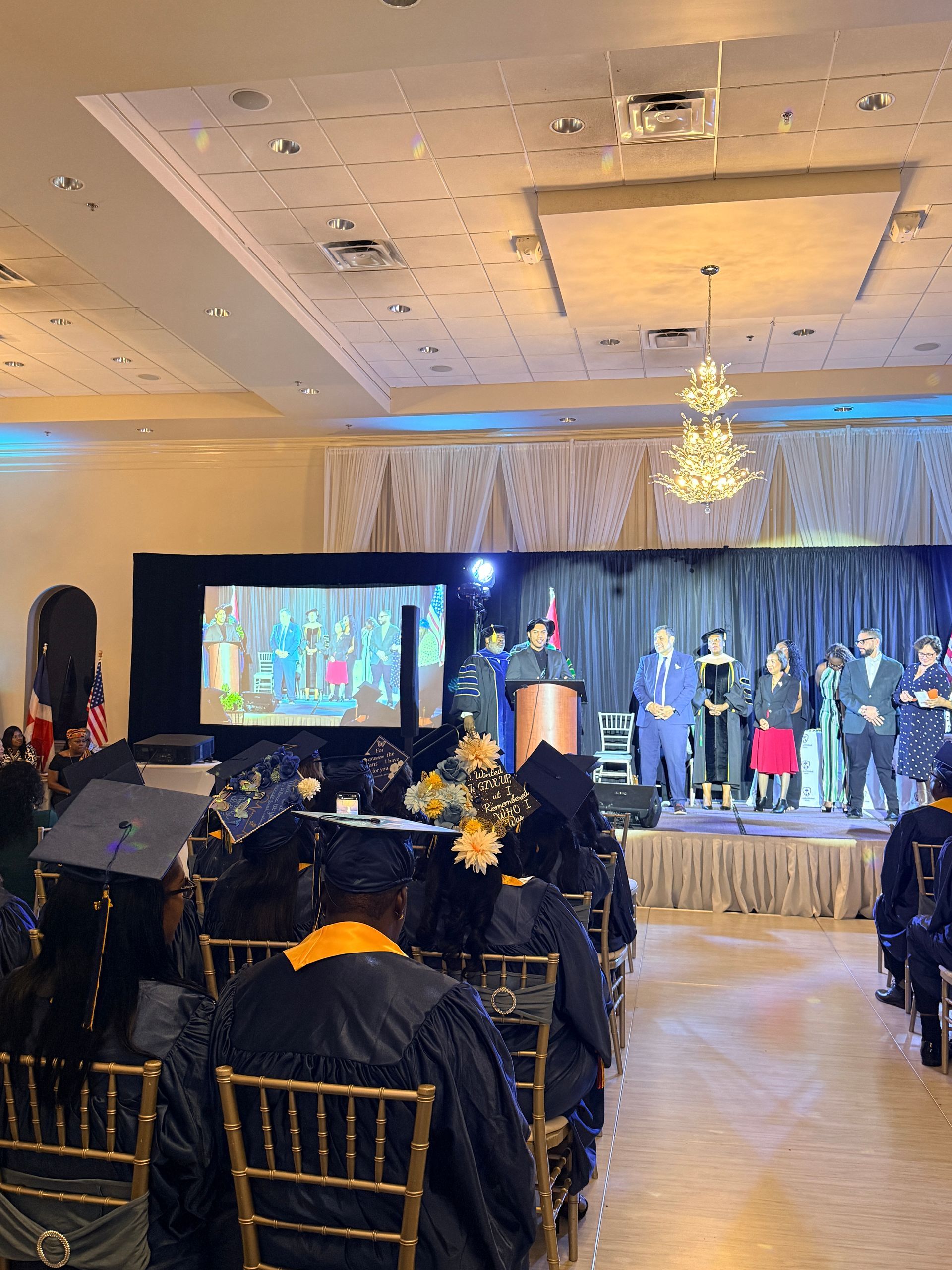 Graduates in caps and gowns sit at a ceremony in a banquet hall, facing a stage with speakers and a projection screen.