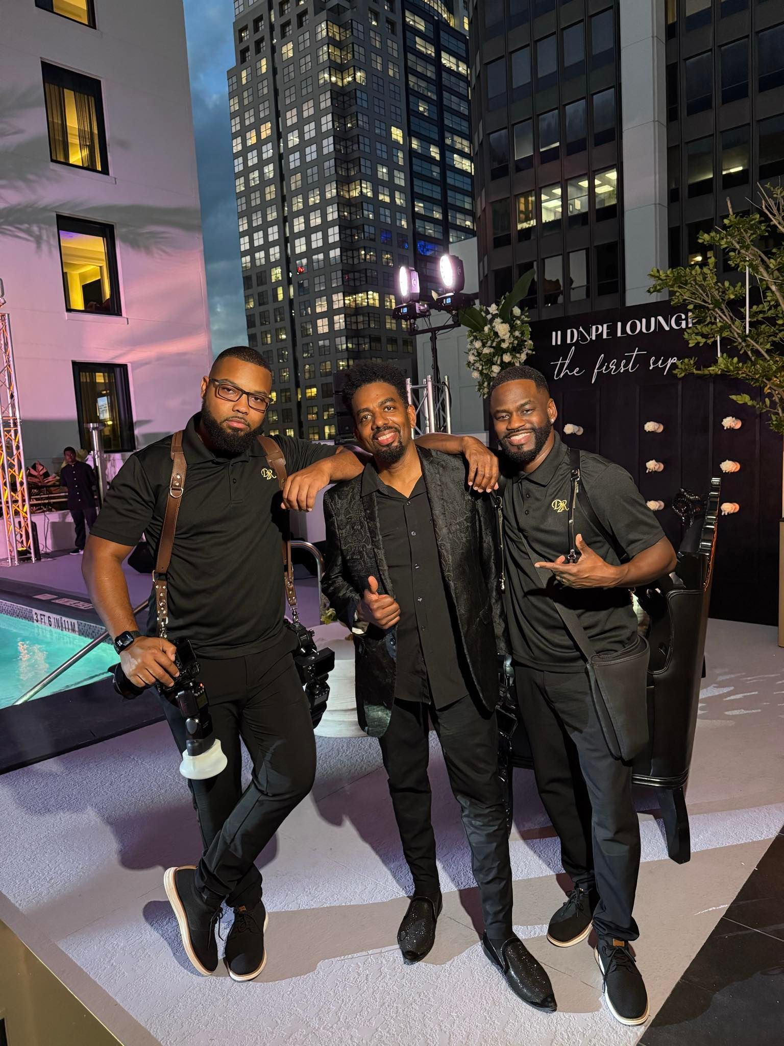 Three men in black outfits and camera equipment pose together on a rooftop at night with city buildings in the background.