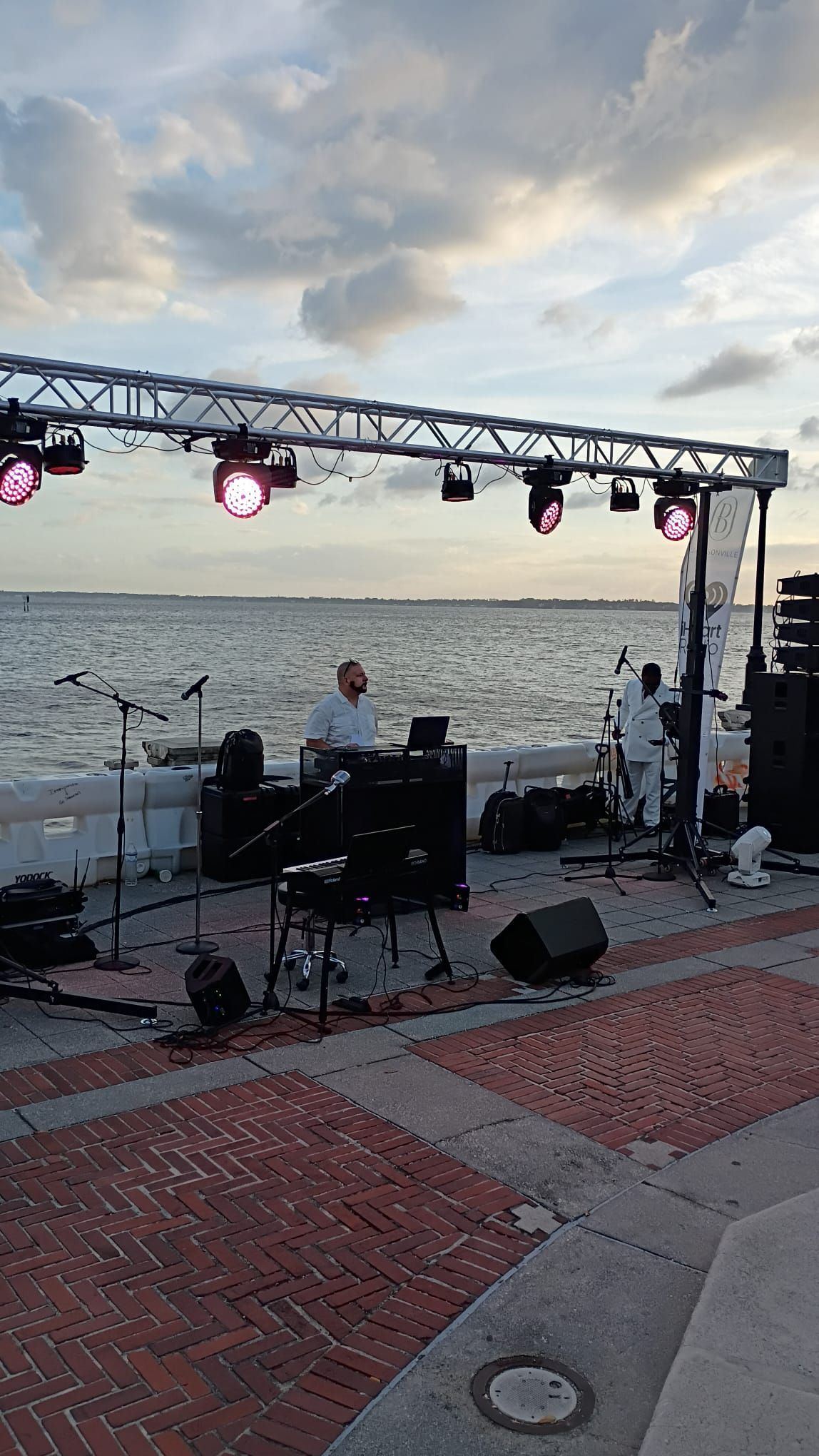 A person performs at an outdoor music setup on a brick patio by the ocean under a cloudy sky with stage lights overhead.