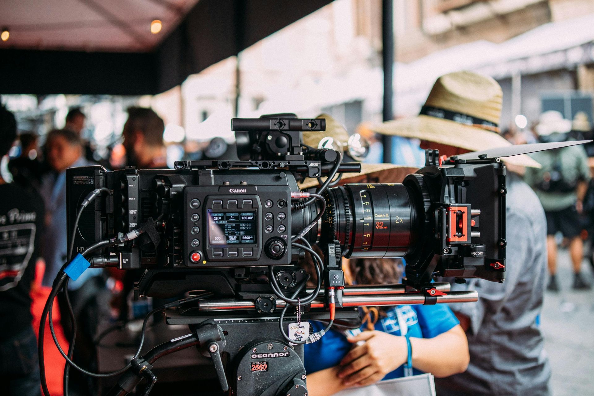 A professional film camera on a tripod, focused on a production set with blurred crew members in the background.