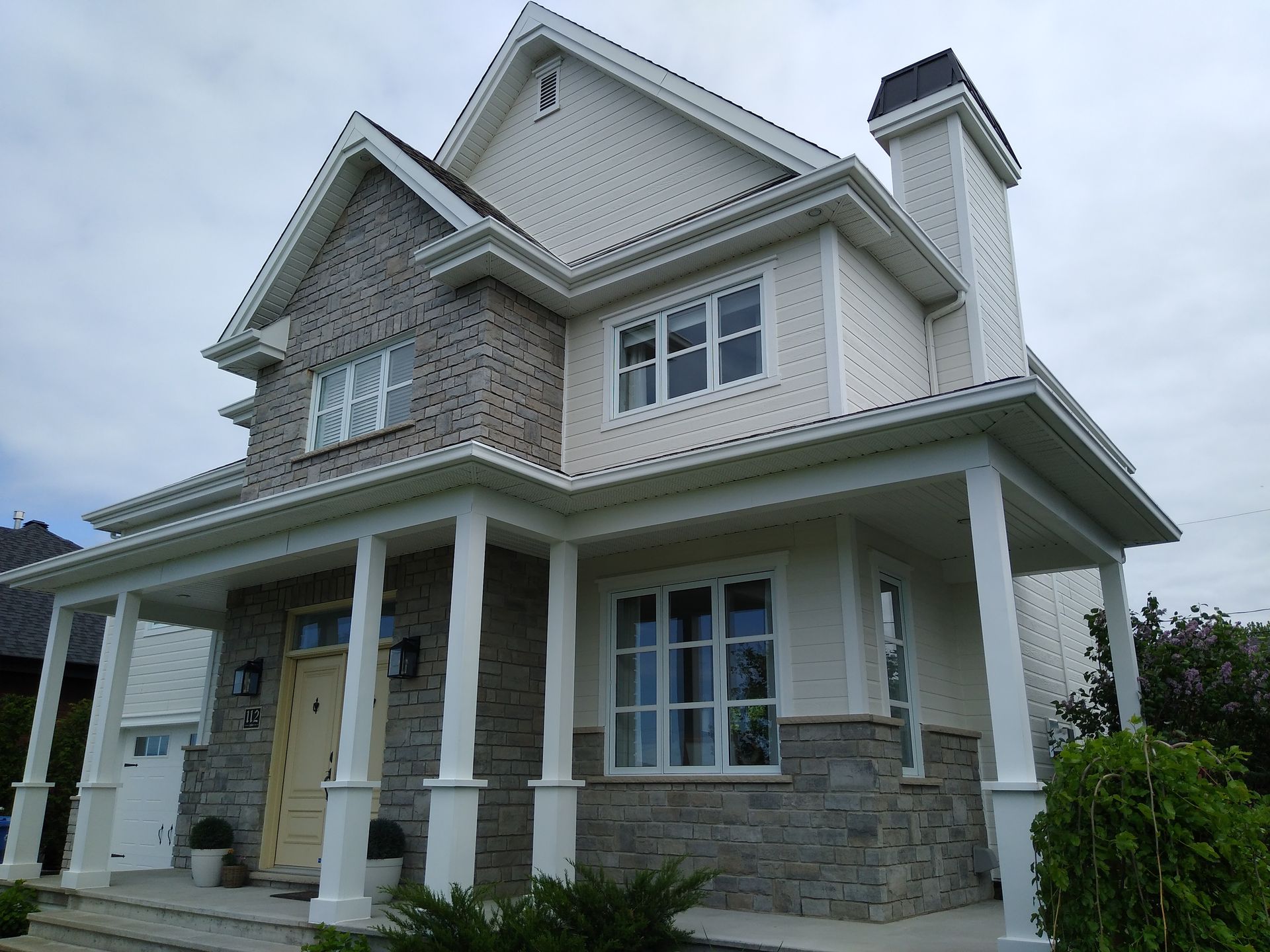 Maison à deux étages avec façade en pierre et bardage clair, porche blanc à colonnes, sous un ciel nuageux.