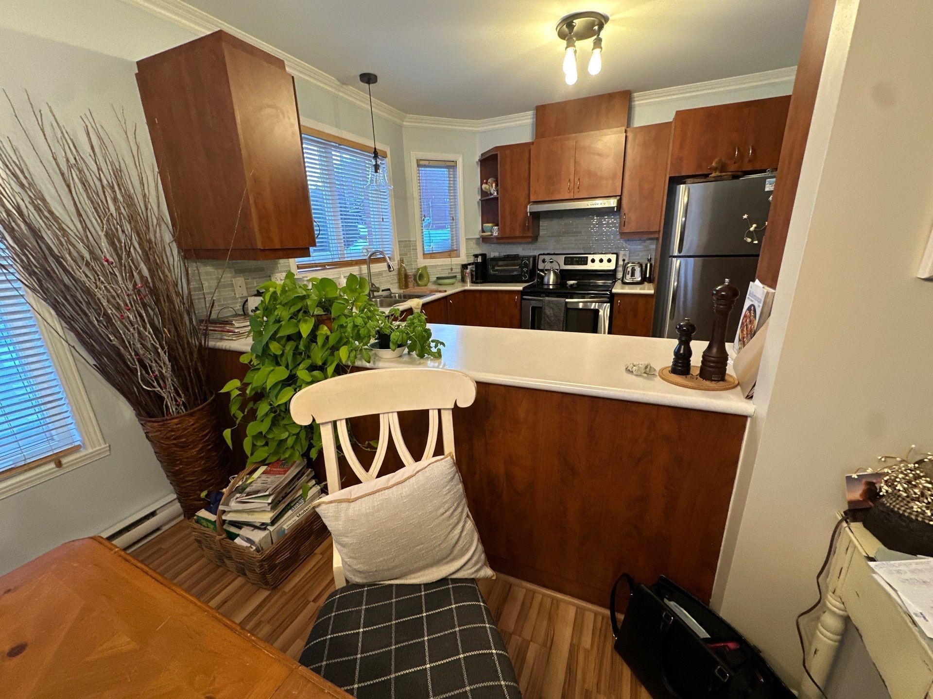 Cuisine avec placards en bois, plan de travail blanc, appareils électroménagers noirs et coin repas avec une chaise.