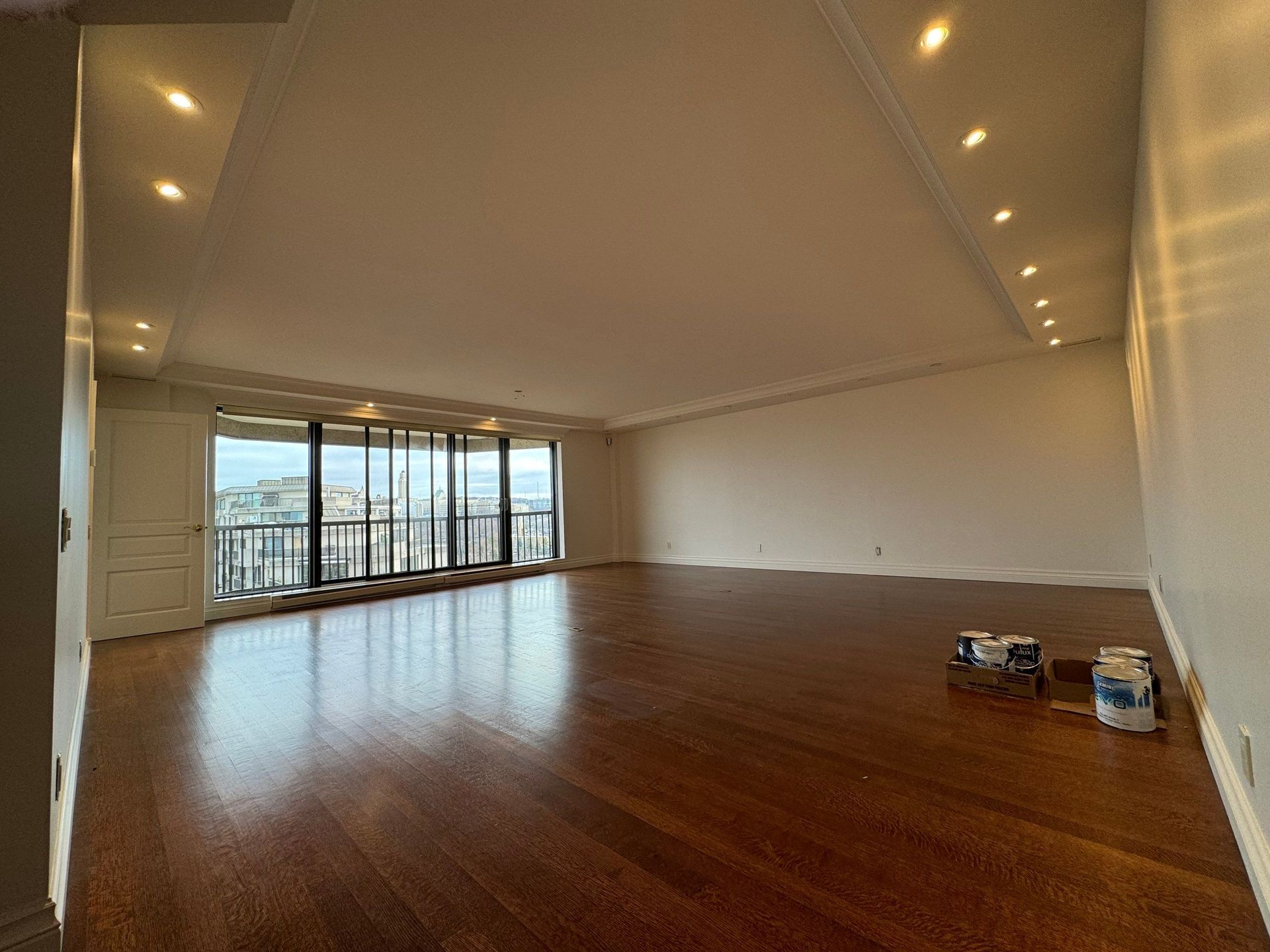 Pièce vide avec parquet, grandes fenêtres, éclairage encastré et vue sur la ville.
