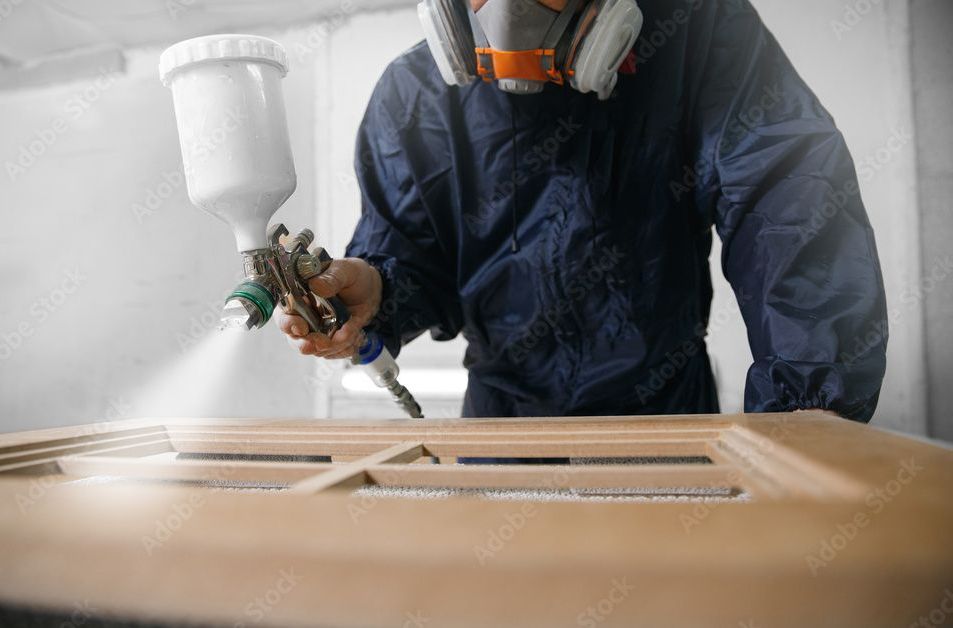 Une personne en équipement de protection peint à la bombe une porte en bois dans un atelier.