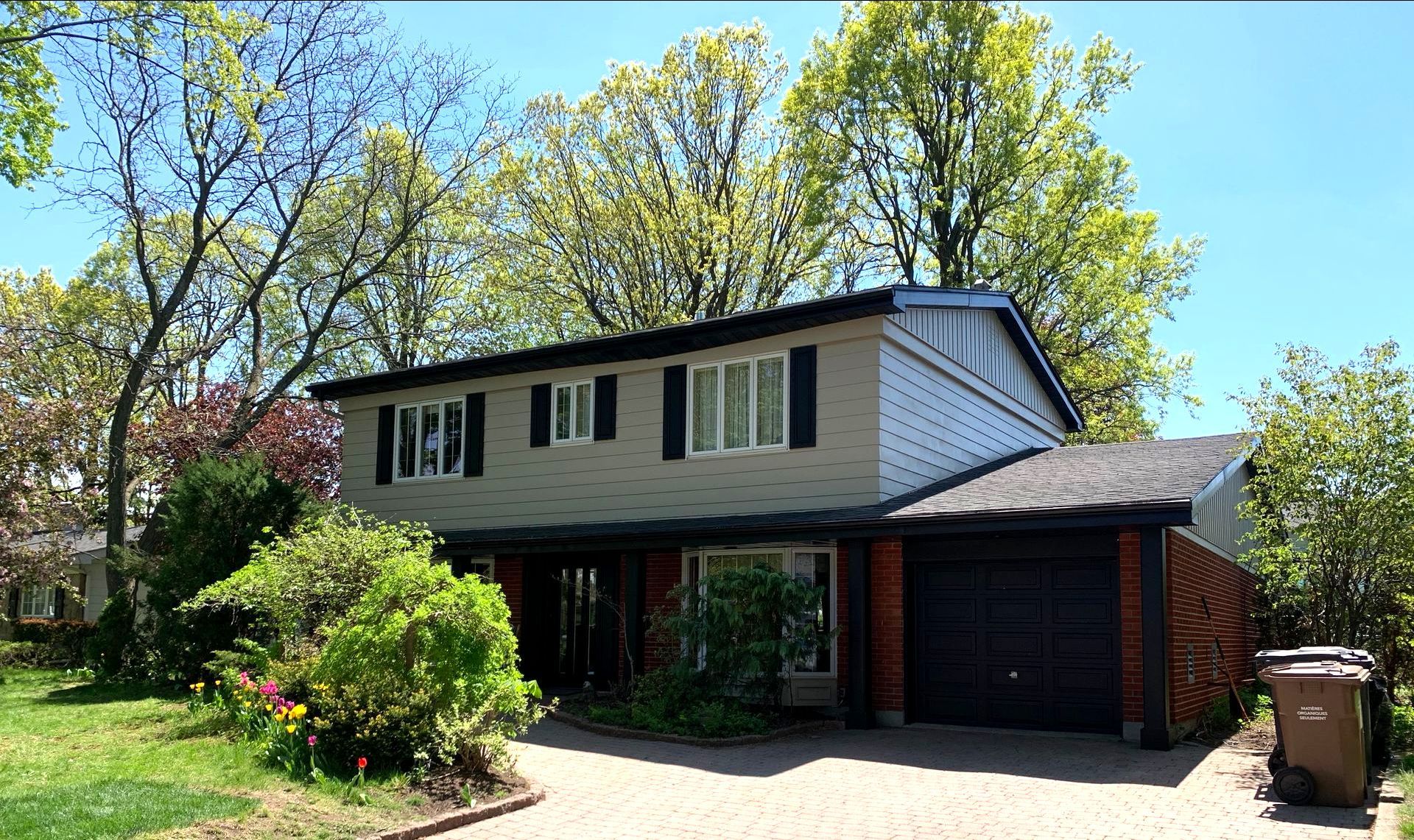 Maison à deux étages avec revêtement gris, volets noirs, garage attenant et arbres matures par une journée ensoleillée.