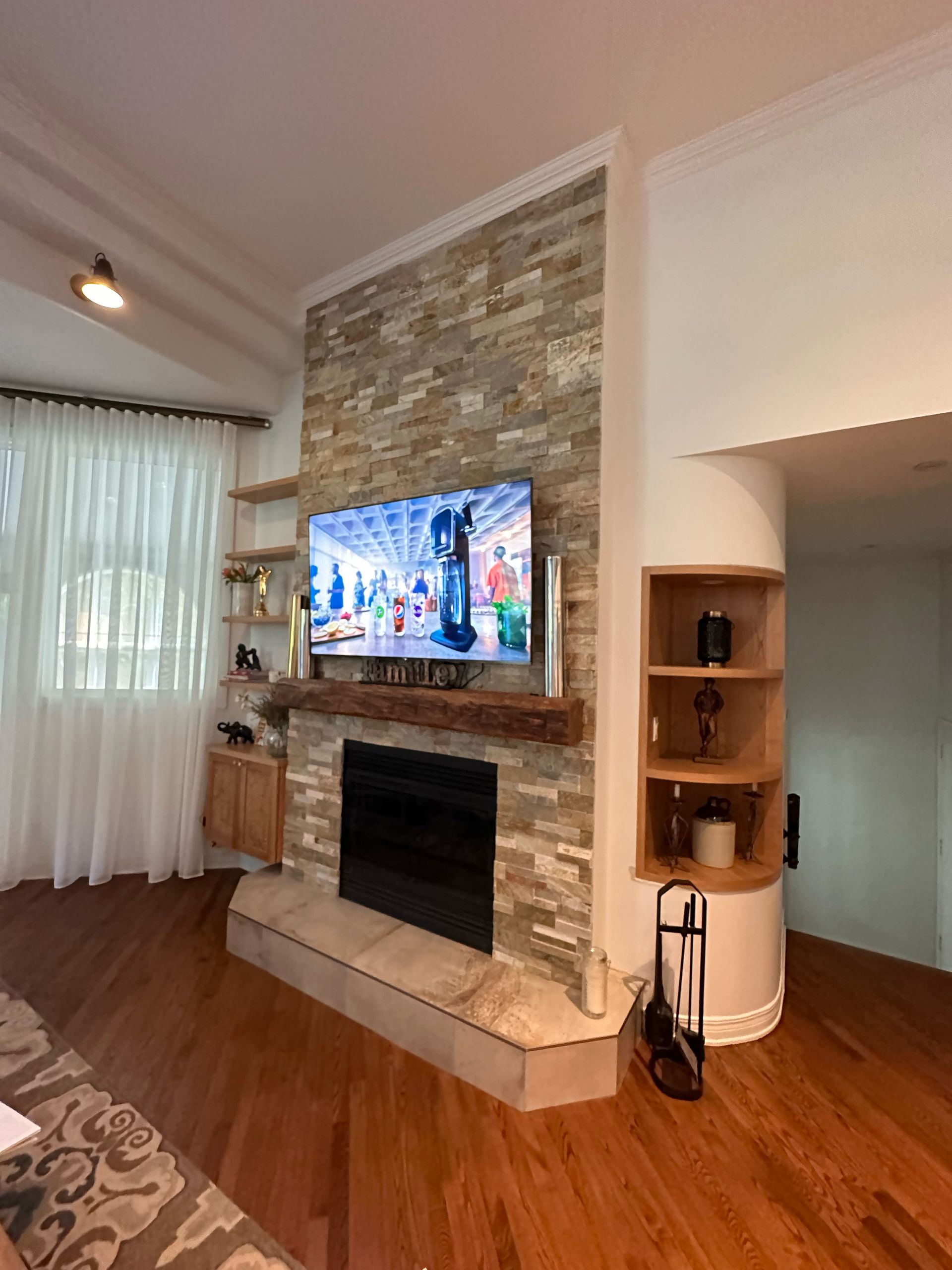 Salon avec cheminée, télévision et étagères encastrées dans un mur en pierre. Parquet et rideaux blancs.